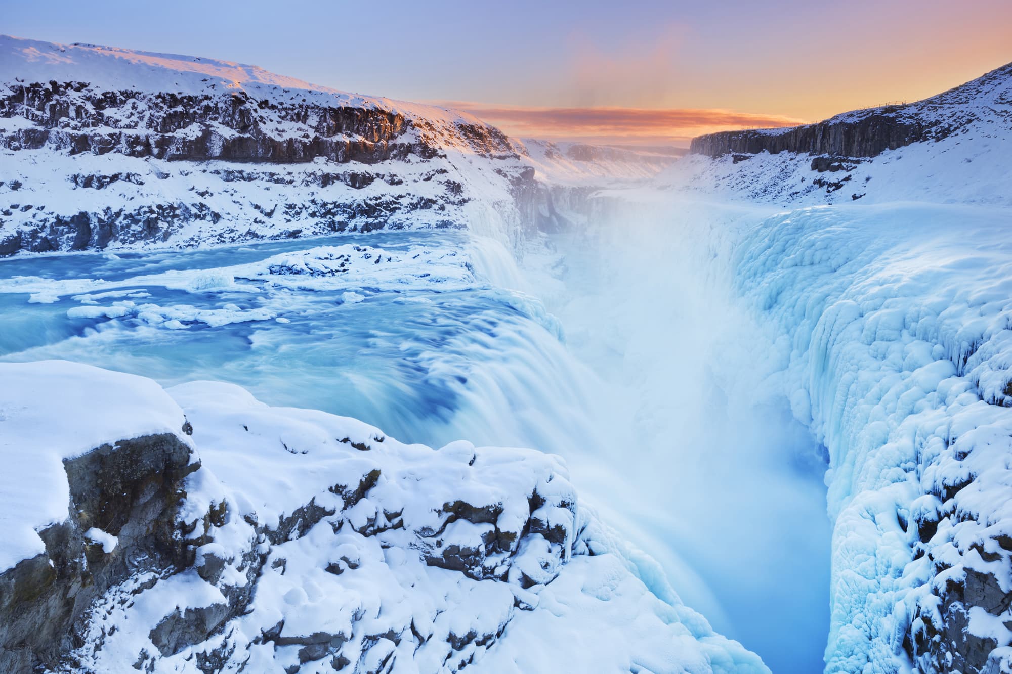 A wide snowy waterfall drops into a frozen canyon at sunrise, with blue water, mist, and orange light on the horizon.