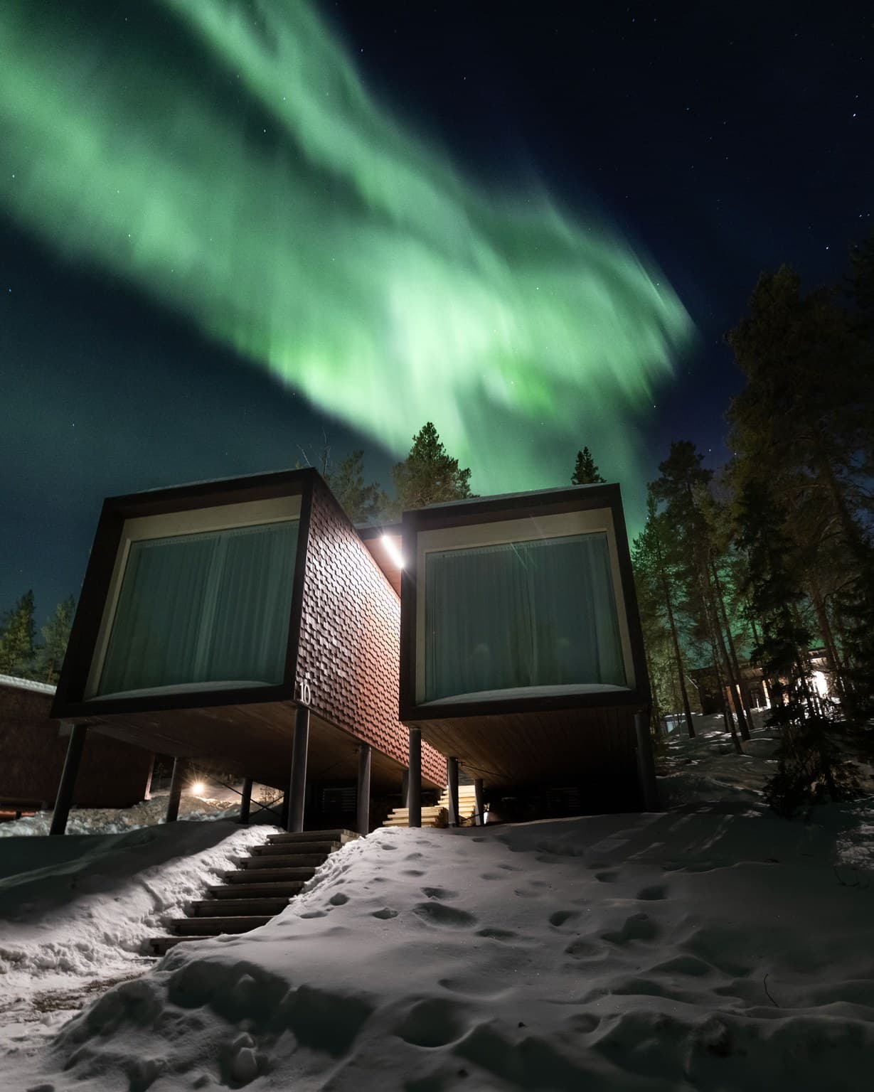 Modern cabin-style rooms on stilts stand in a snowy forest under bright green northern lights at night.