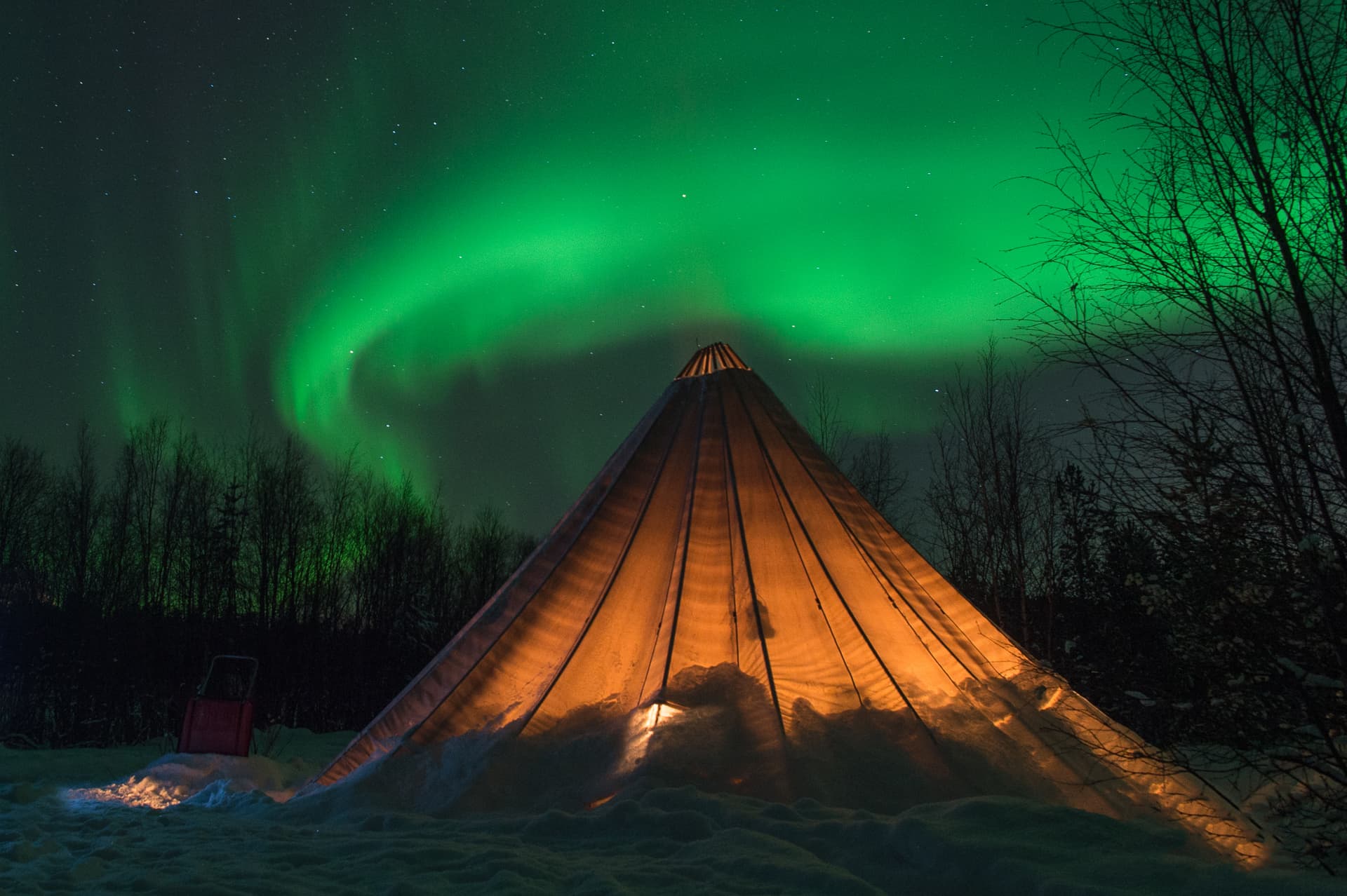 A glowing conical tent stands in a snowy landscape beneath vivid green northern lights in a starry night sky.