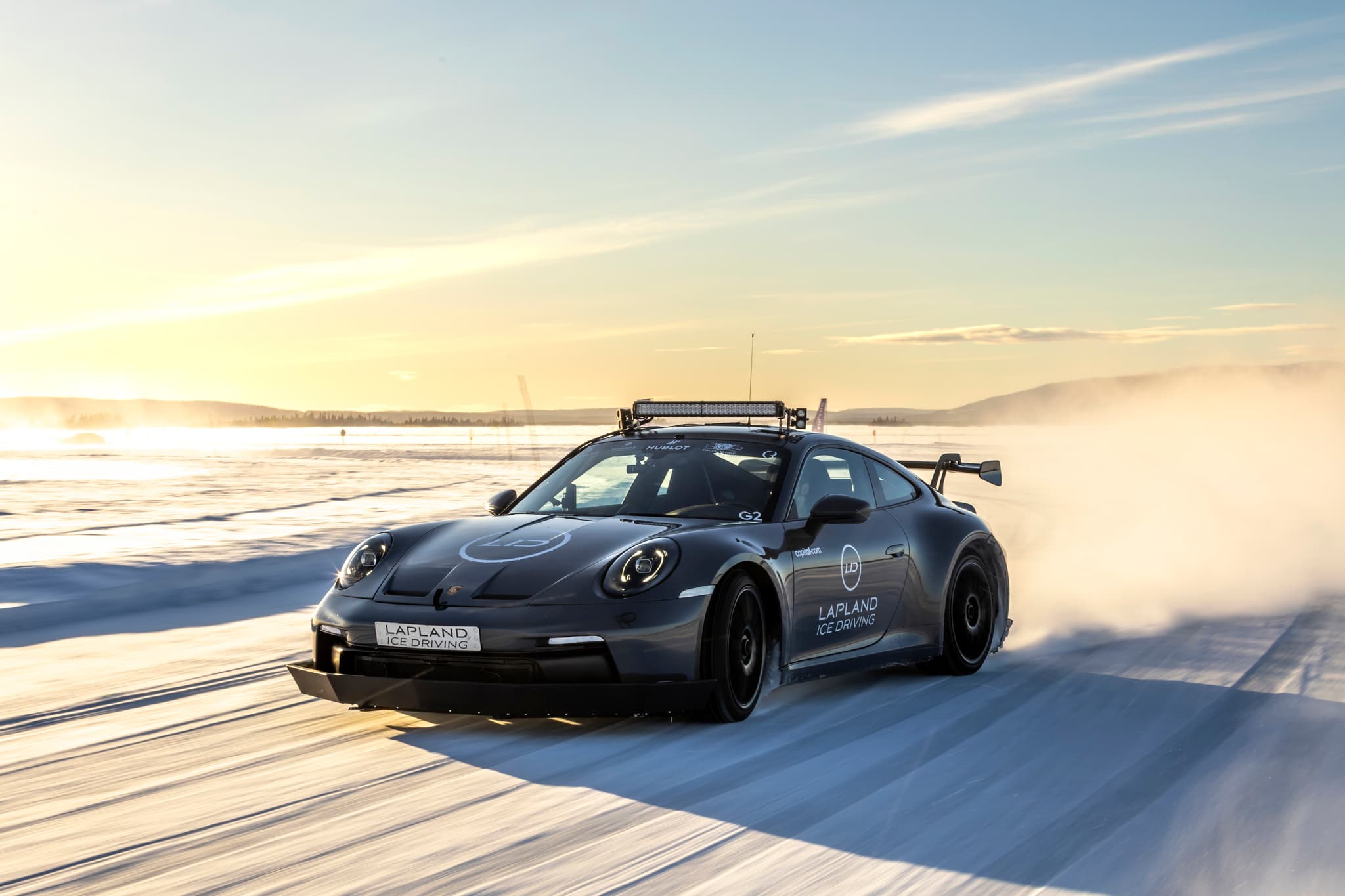 A black sports car drifts across an icy track at sunset, throwing up a cloud of snow.