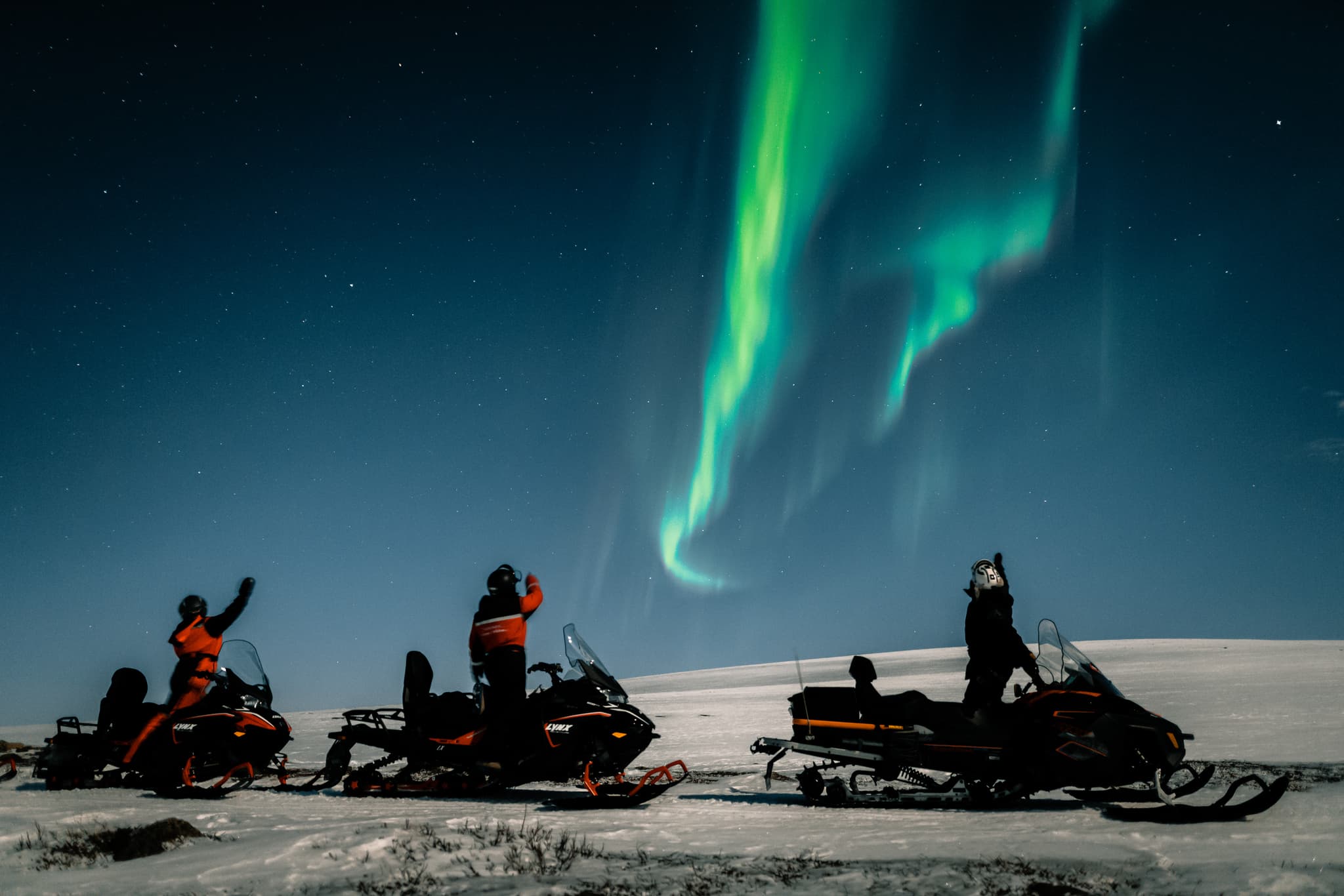 Snowmobiles on a snowy landscape under vivid green aurora borealis with starry night sky.
