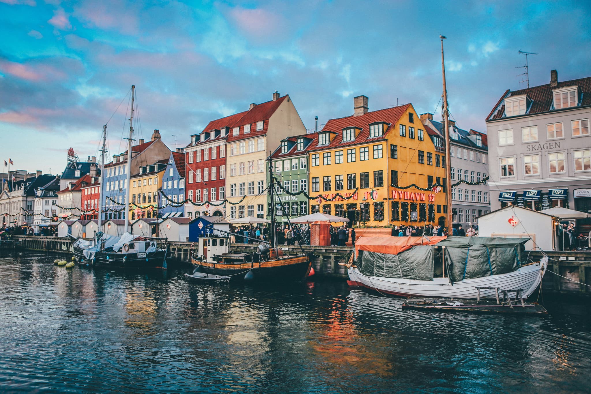 Harbor view with moored sailboats and colorful waterfront buildings with red tile roofs under a blue sky with clouds.