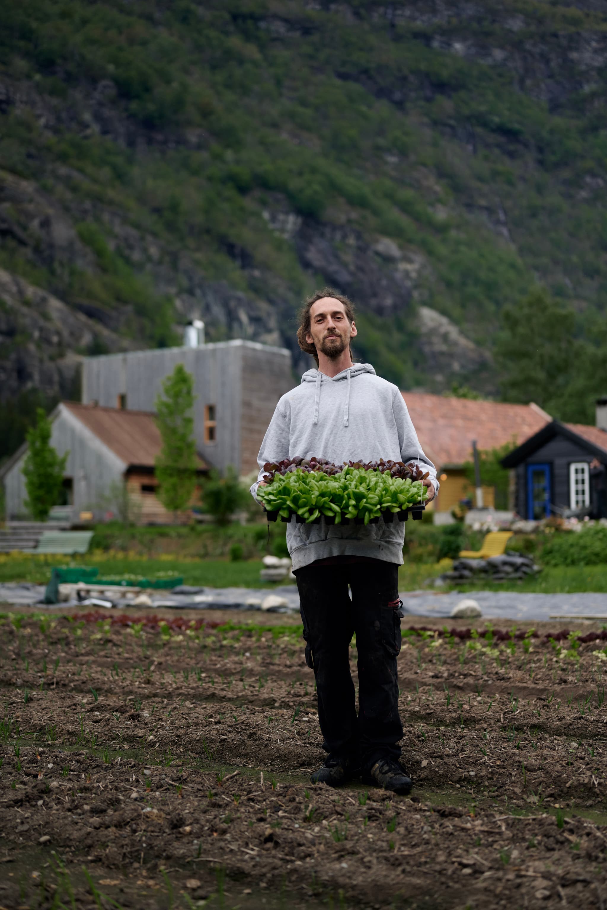 A man in a gray hoodie stands in a vegetable garden holding trays of seedlings with mountains and houses in the background.