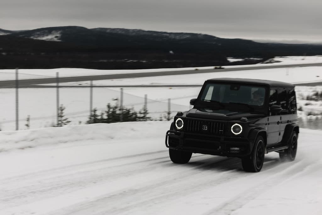 A black SUV drives along a snowy road with headlights on in a winter landscape.