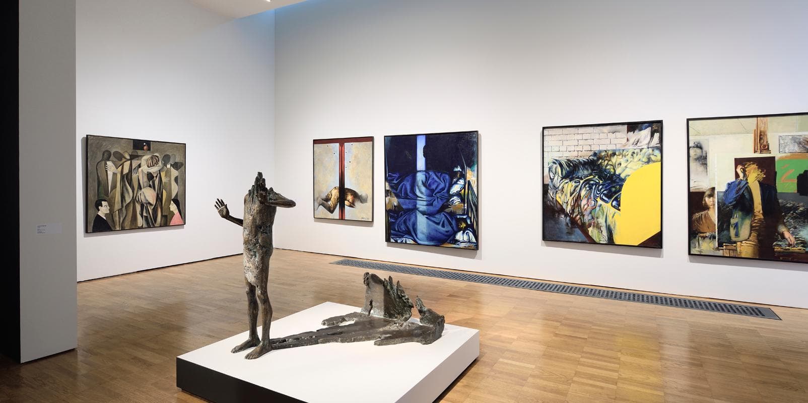 A modern art gallery room displays several large paintings on white walls and a bronze sculpture on a white pedestal in the center of the wooden floor.