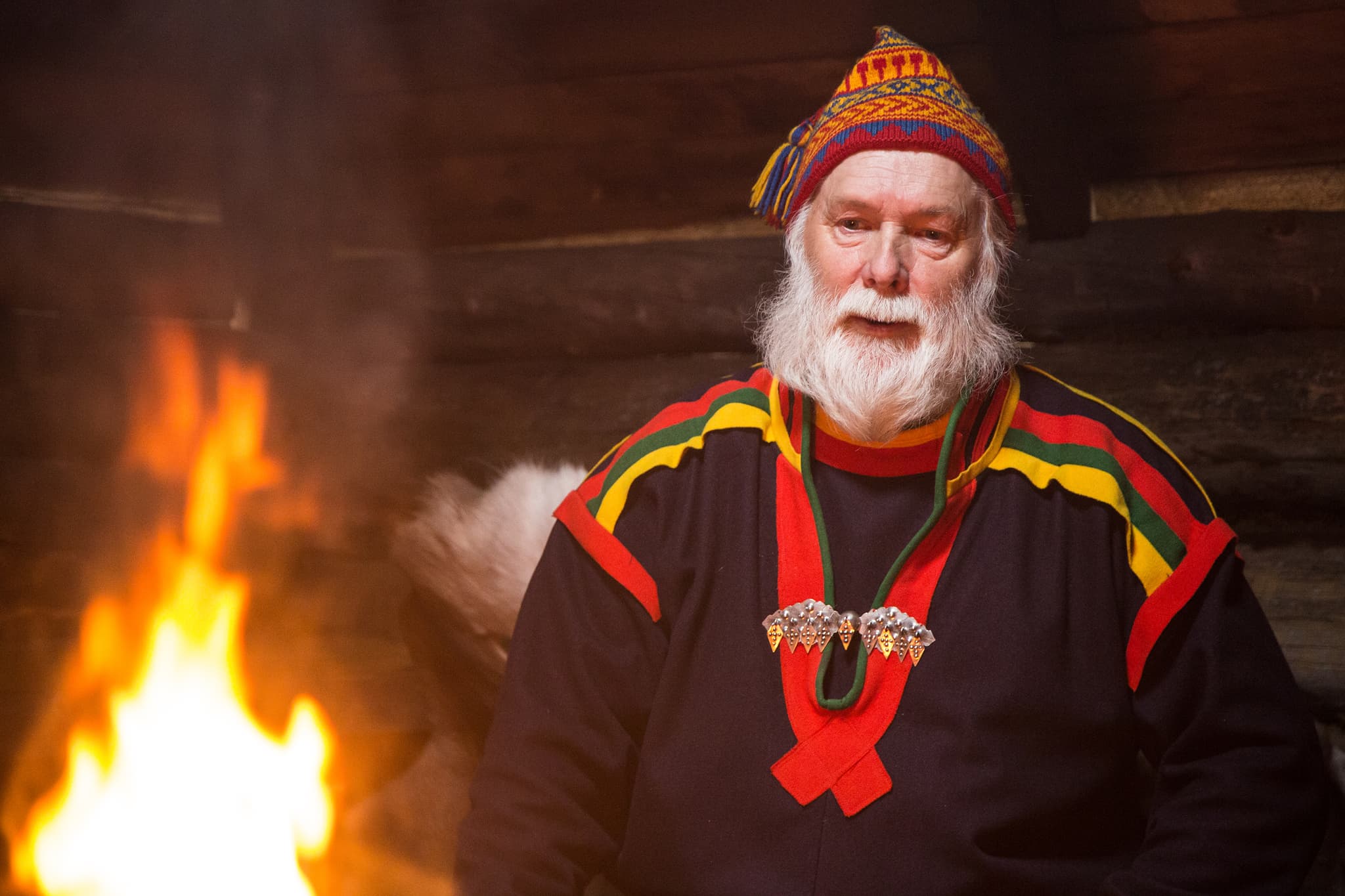 An elderly man in colorful traditional Sami dress and knitted hat stands indoors beside a fire.