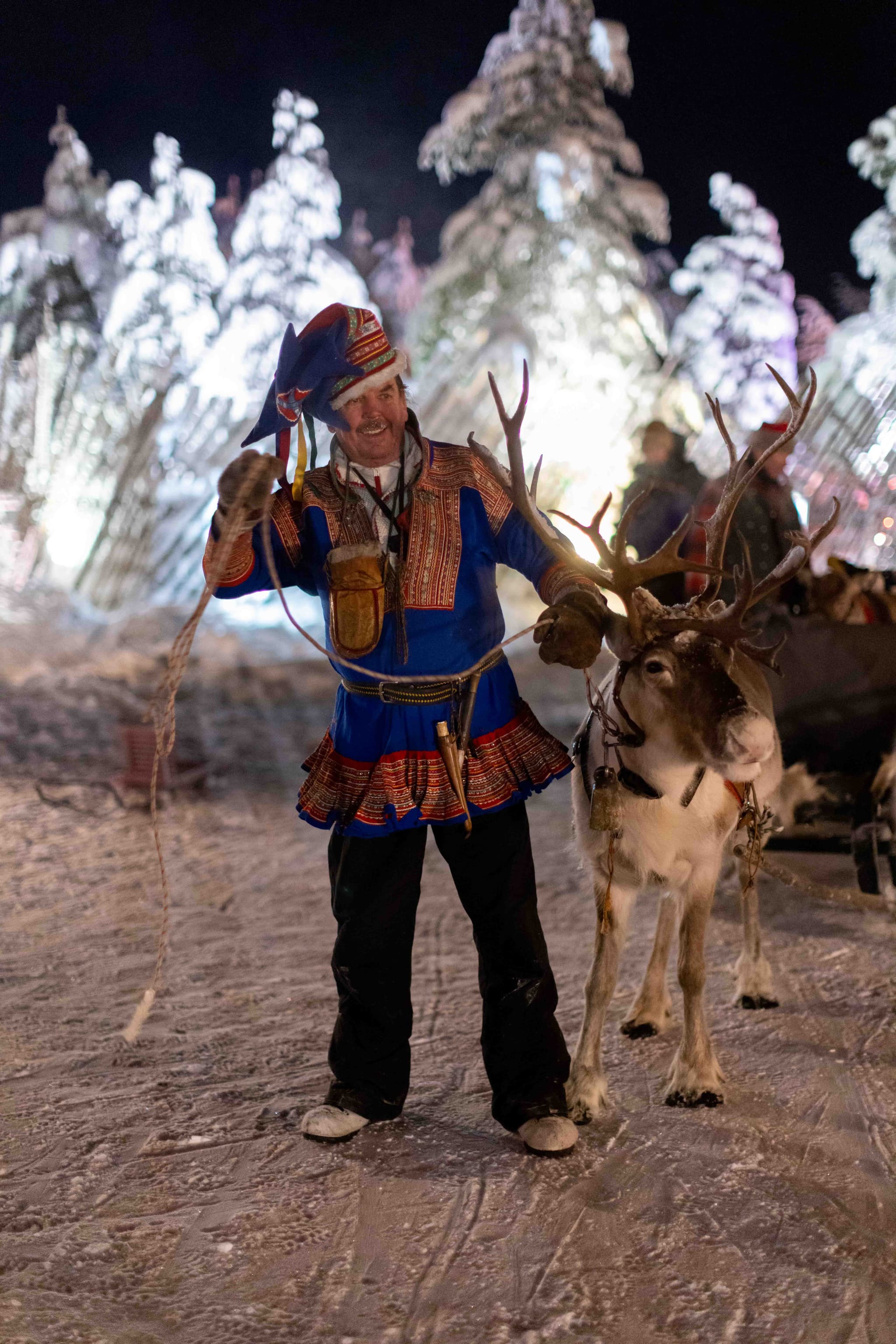 Smiling reindeer handler in traditional Sami-style outfit stands on snowy ground beside a reindeer with antlers, with snowy trees and onlookers in the night background.