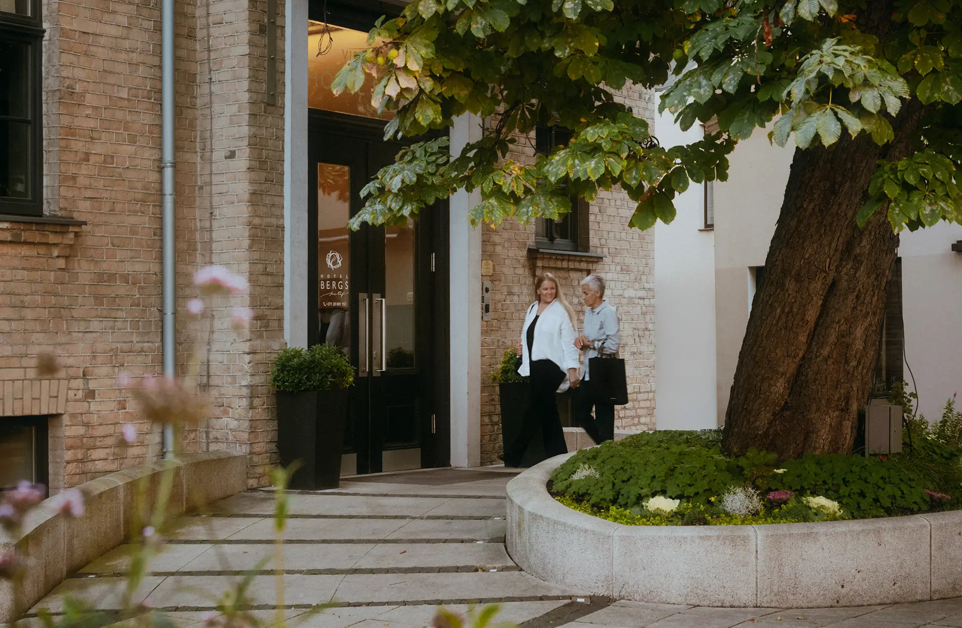Two women walk past a brick building entrance shaded by a large tree and surrounded by planted greenery.