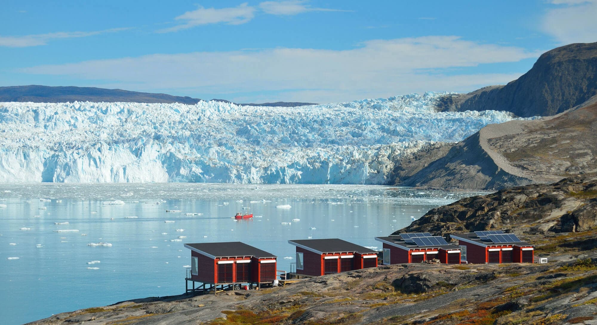 A wide glacier fills a fjord beside a rocky shoreline with several small red cabins in the foreground and a tiny boat on the calm water.