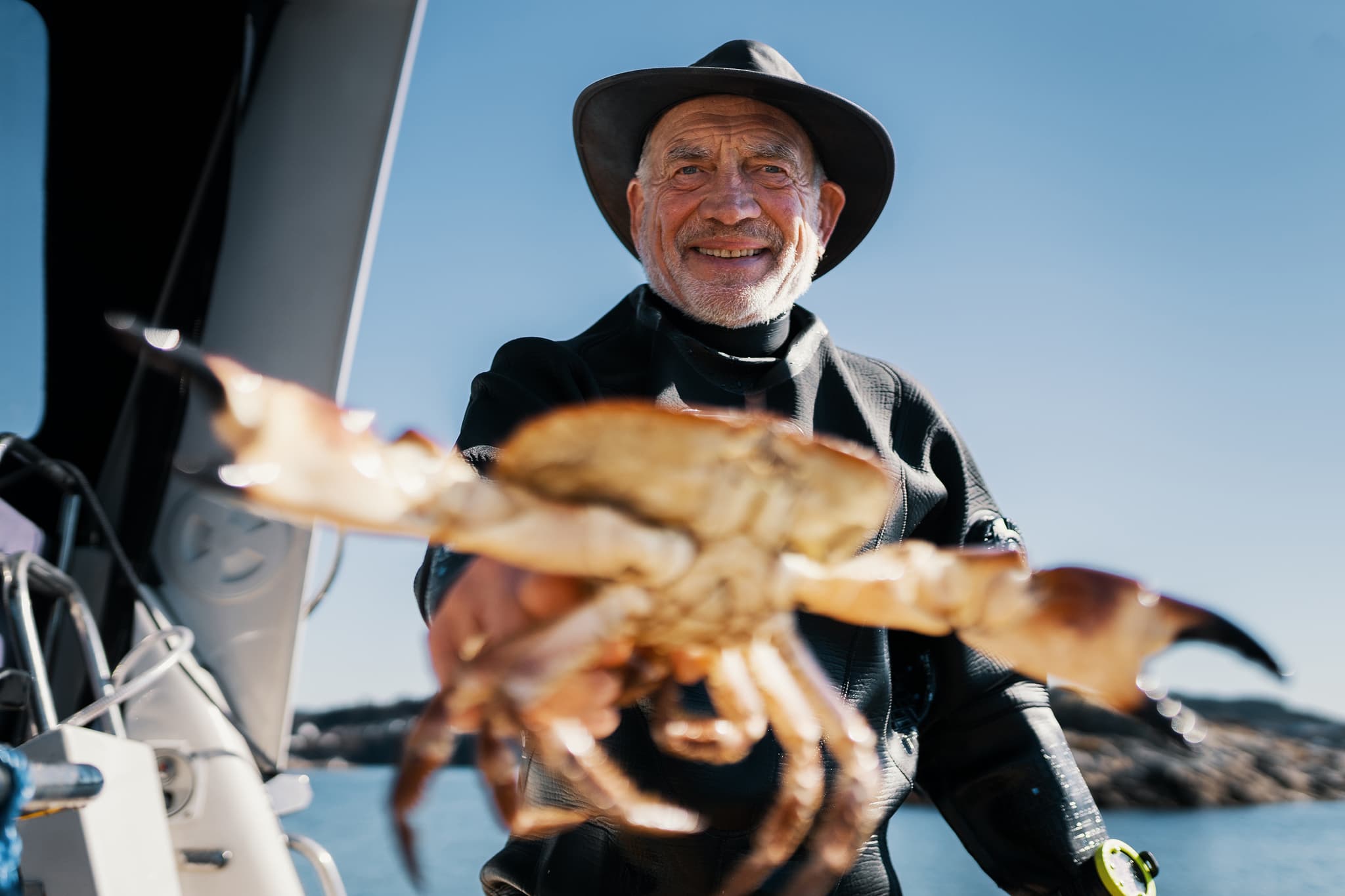 An older man in a black wetsuit and wide-brimmed hat holds a crab toward the camera on a boat with calm water and rocky shoreline behind him.