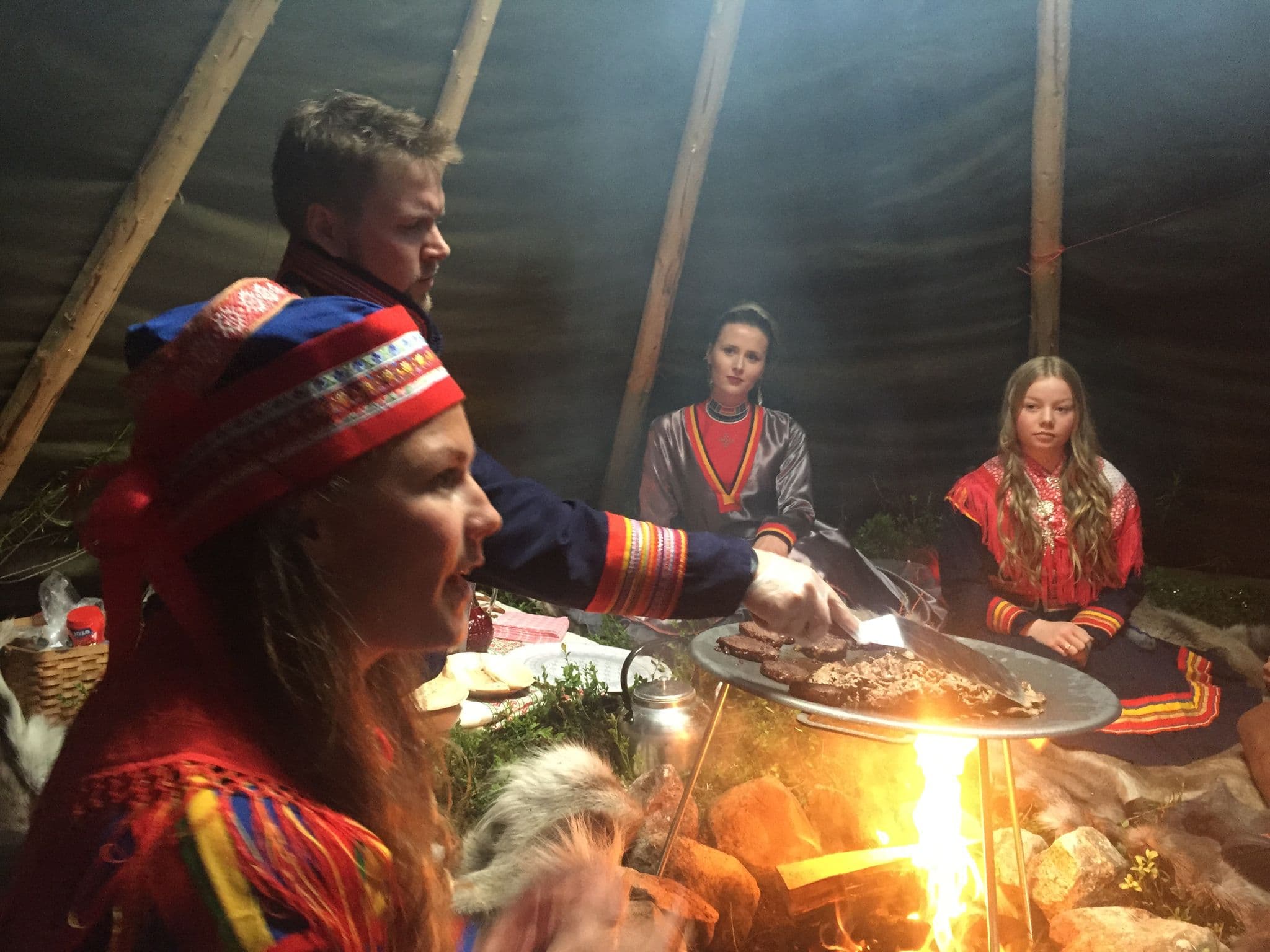 A group of people in colorful traditional clothing gathers around an open fire while food cooks on a grill inside a tent-like shelter.