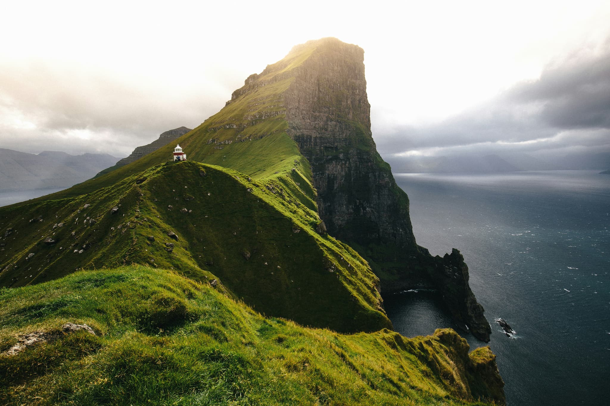 A small lighthouse stands on a grassy ridge beside a dramatic sea cliff overlooking dark ocean water under a cloudy sky.