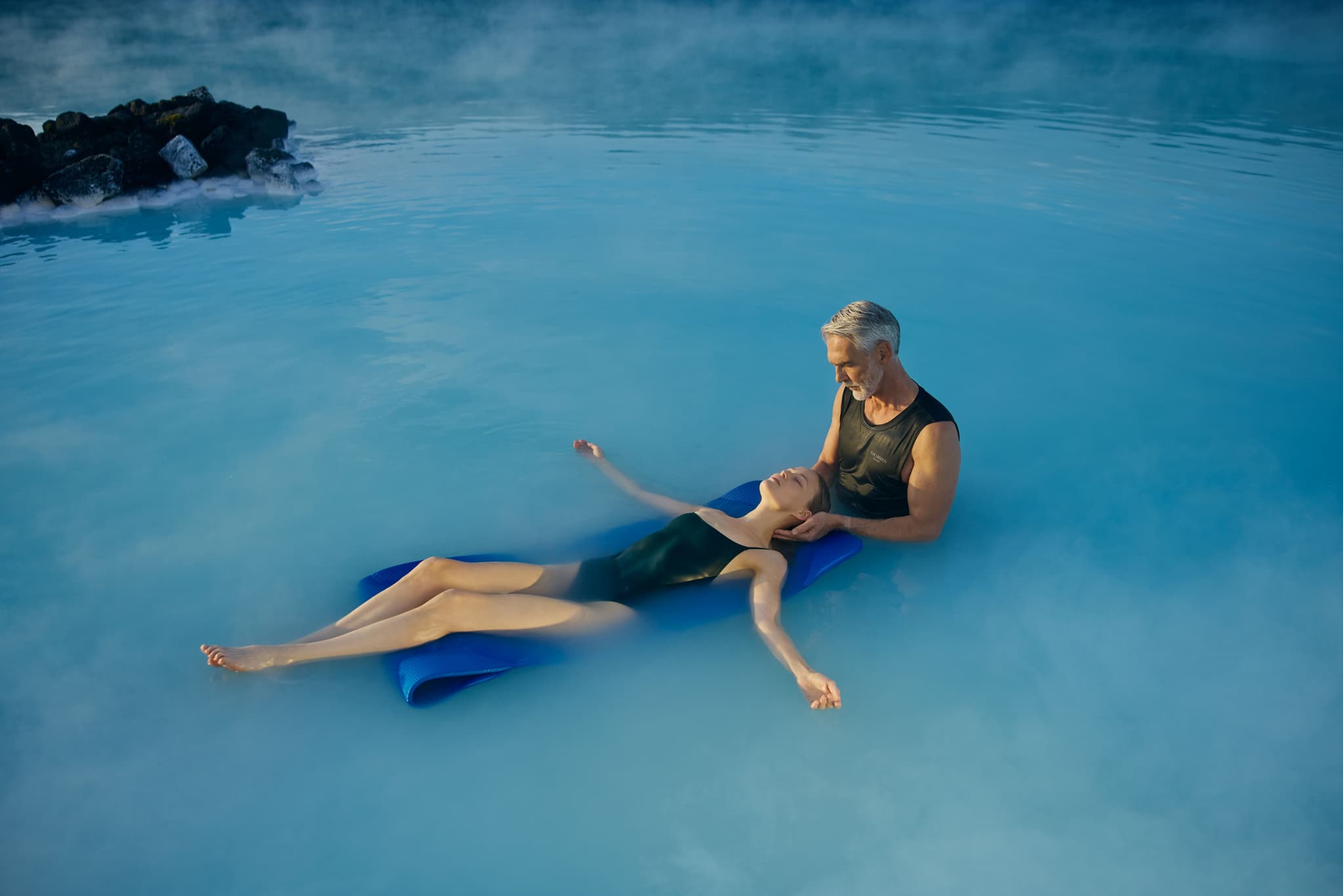 Two people performing a water massage while floating on a blue mat in a calm pool or spa with rocky landscaping in the background.