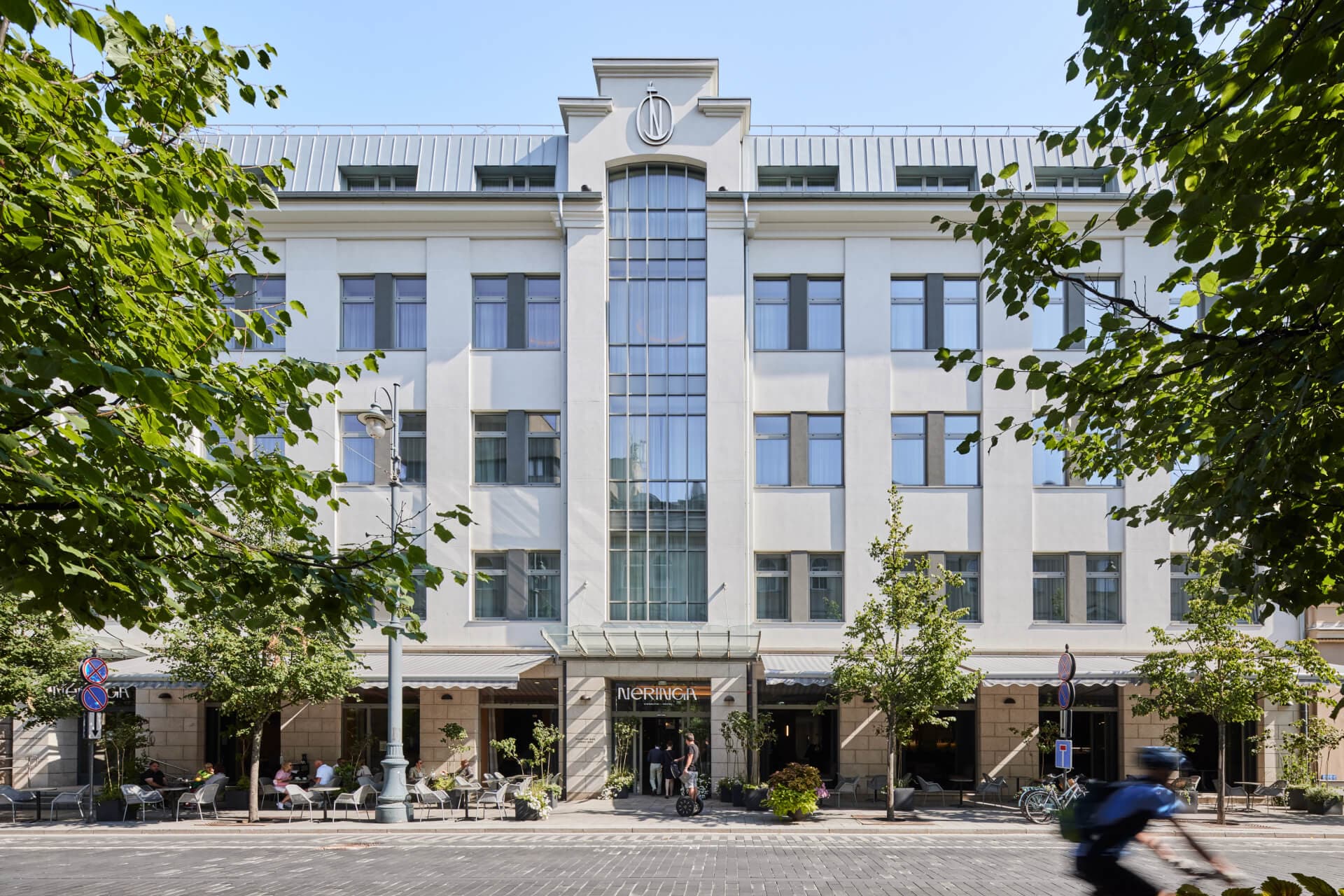A white multi-story hotel facade with a central glass section is framed by leafy trees on a sunny day, with outdoor seating and a cyclist passing in front.