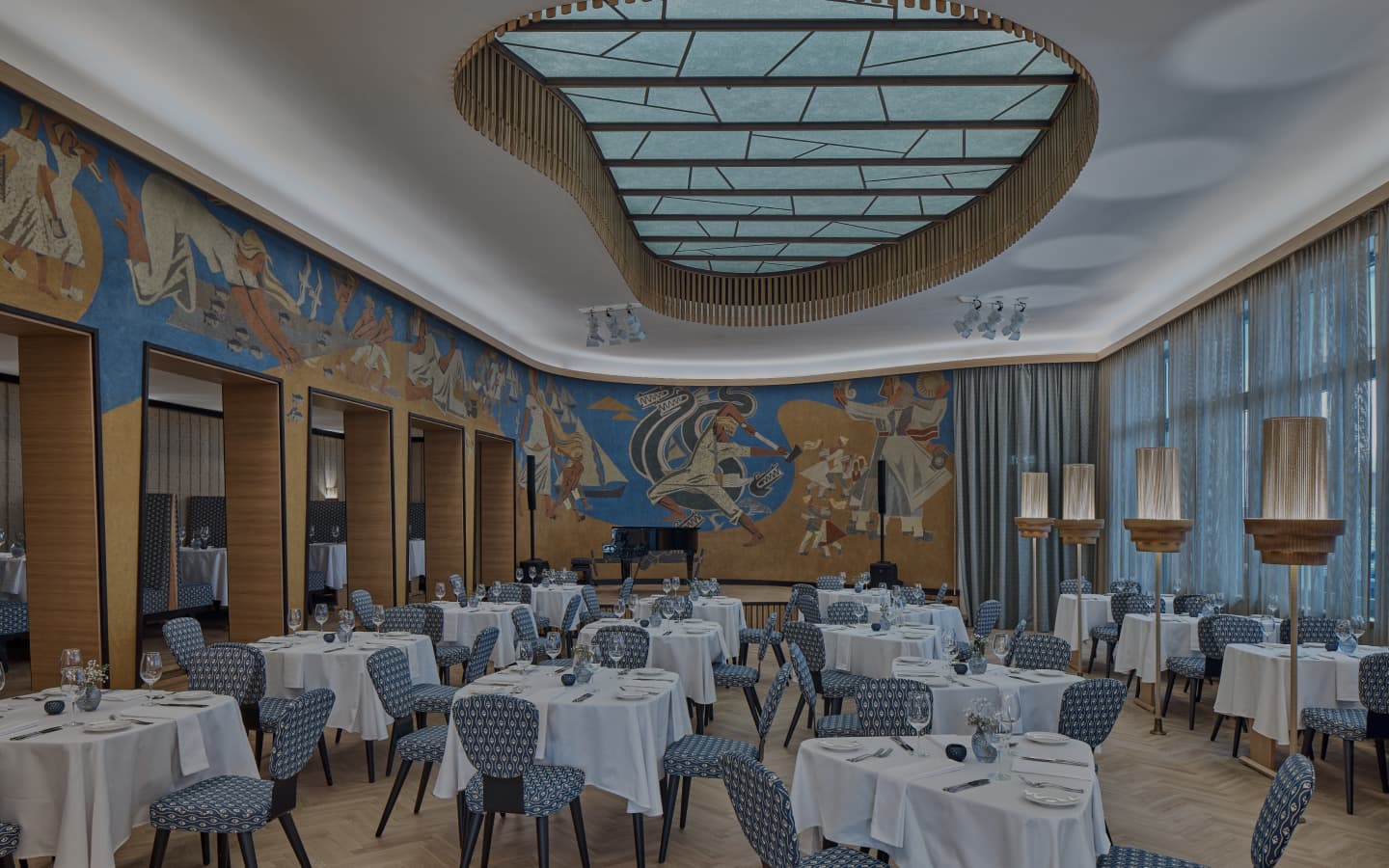 An elegant dining room with round tables set with white tablecloths, patterned blue chairs, and a large mural along the wall.