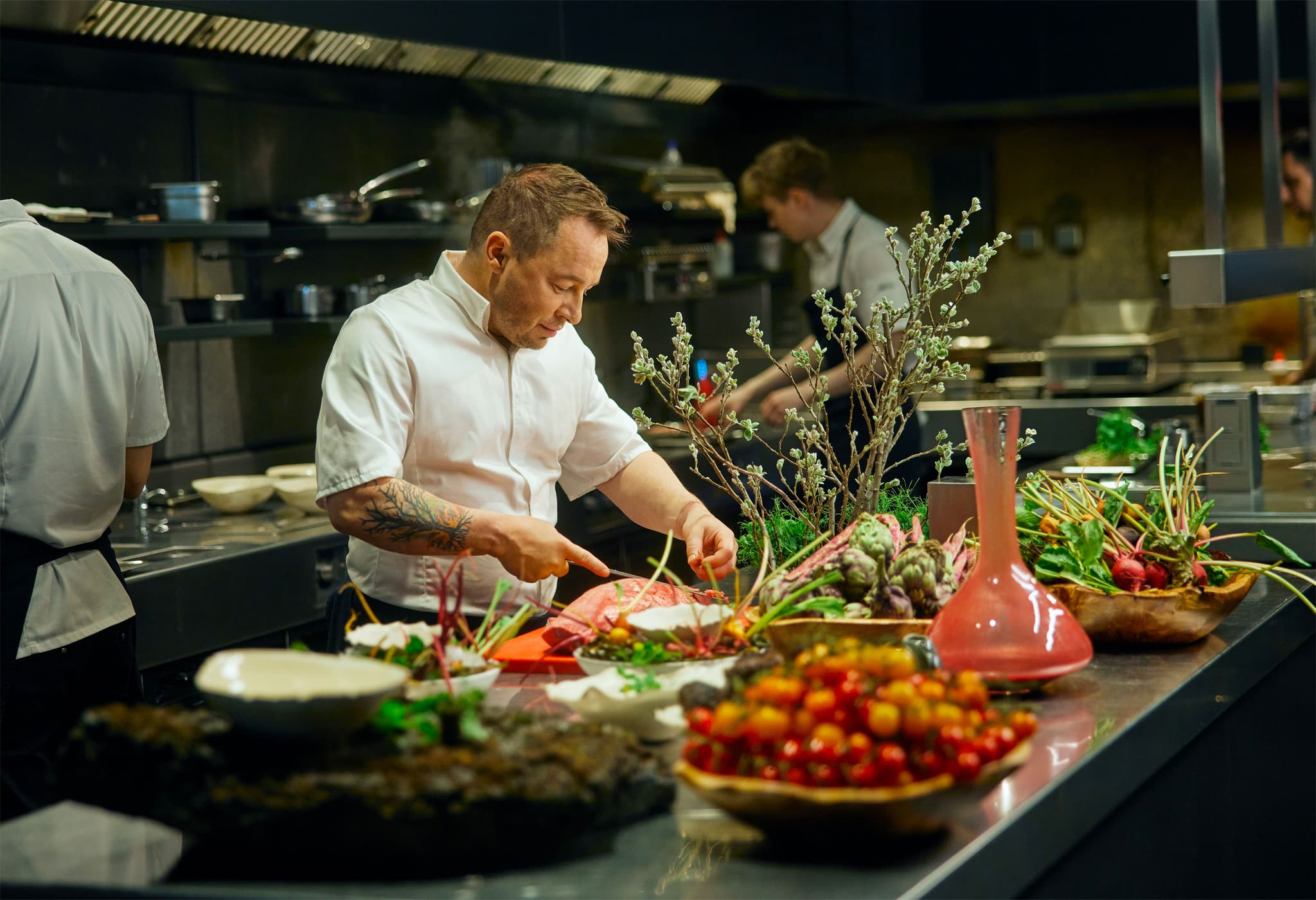 Chef in white coat preparing ingredients in a professional kitchen, with plated vegetables and herbs in the foreground.