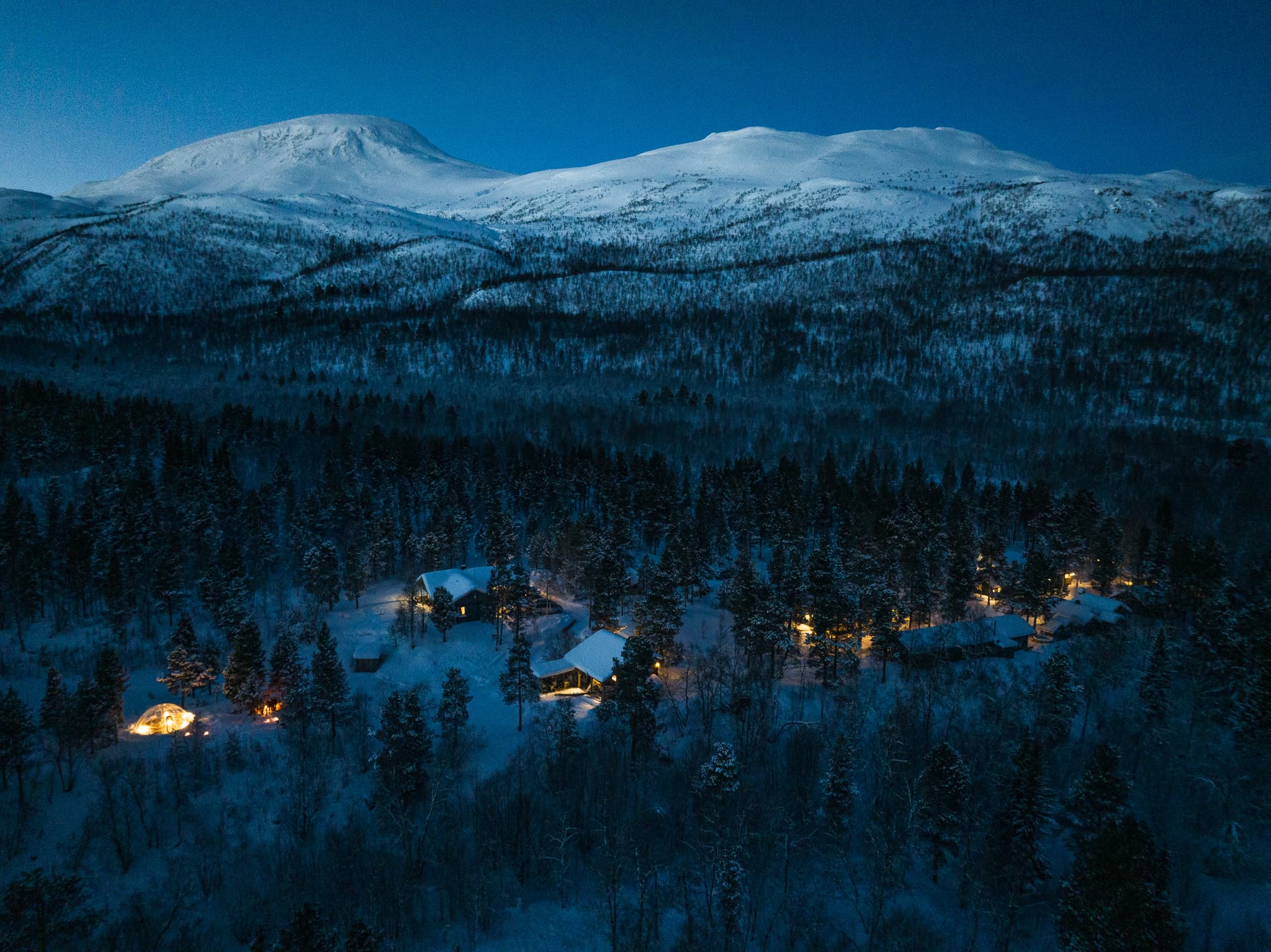 A snowy forest landscape with a small cluster of warmly lit cabins at night beneath a range of snow-covered mountains.