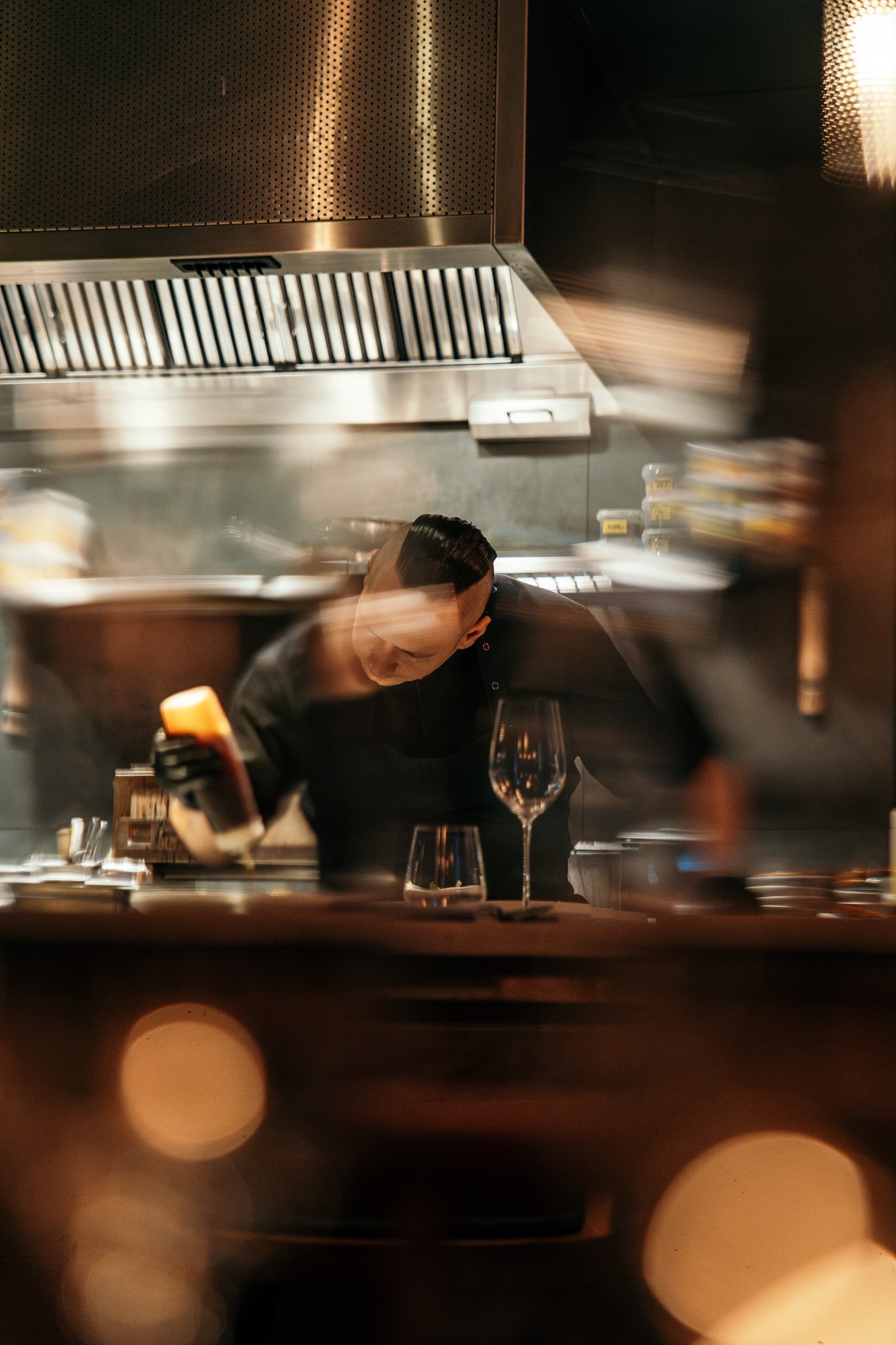 A chef in a dark kitchen is plating food behind a counter with wine glasses in the foreground.