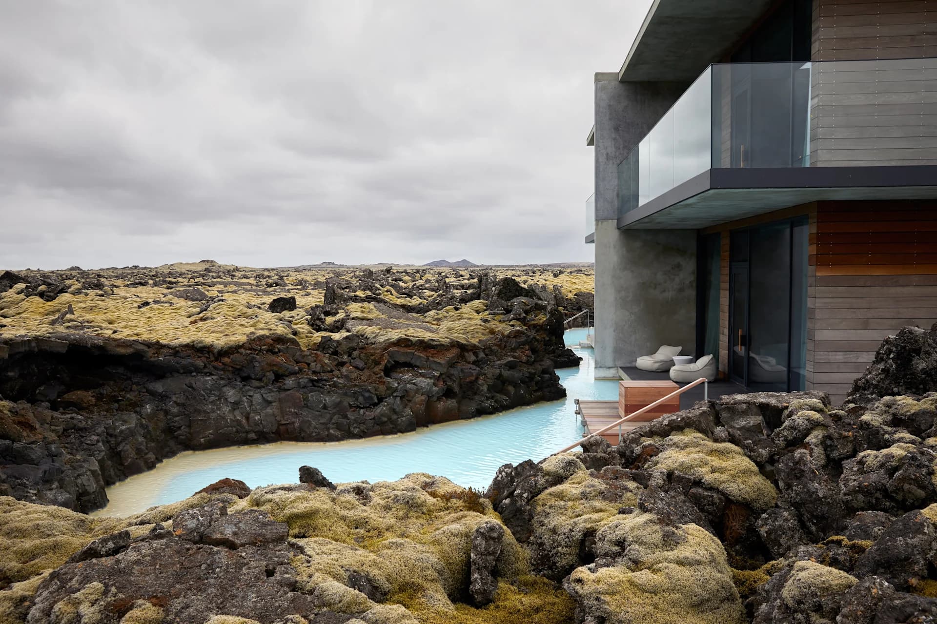A modern hotel building stands beside a pale blue geothermal water channel flowing through a rugged lava field under an overcast sky.