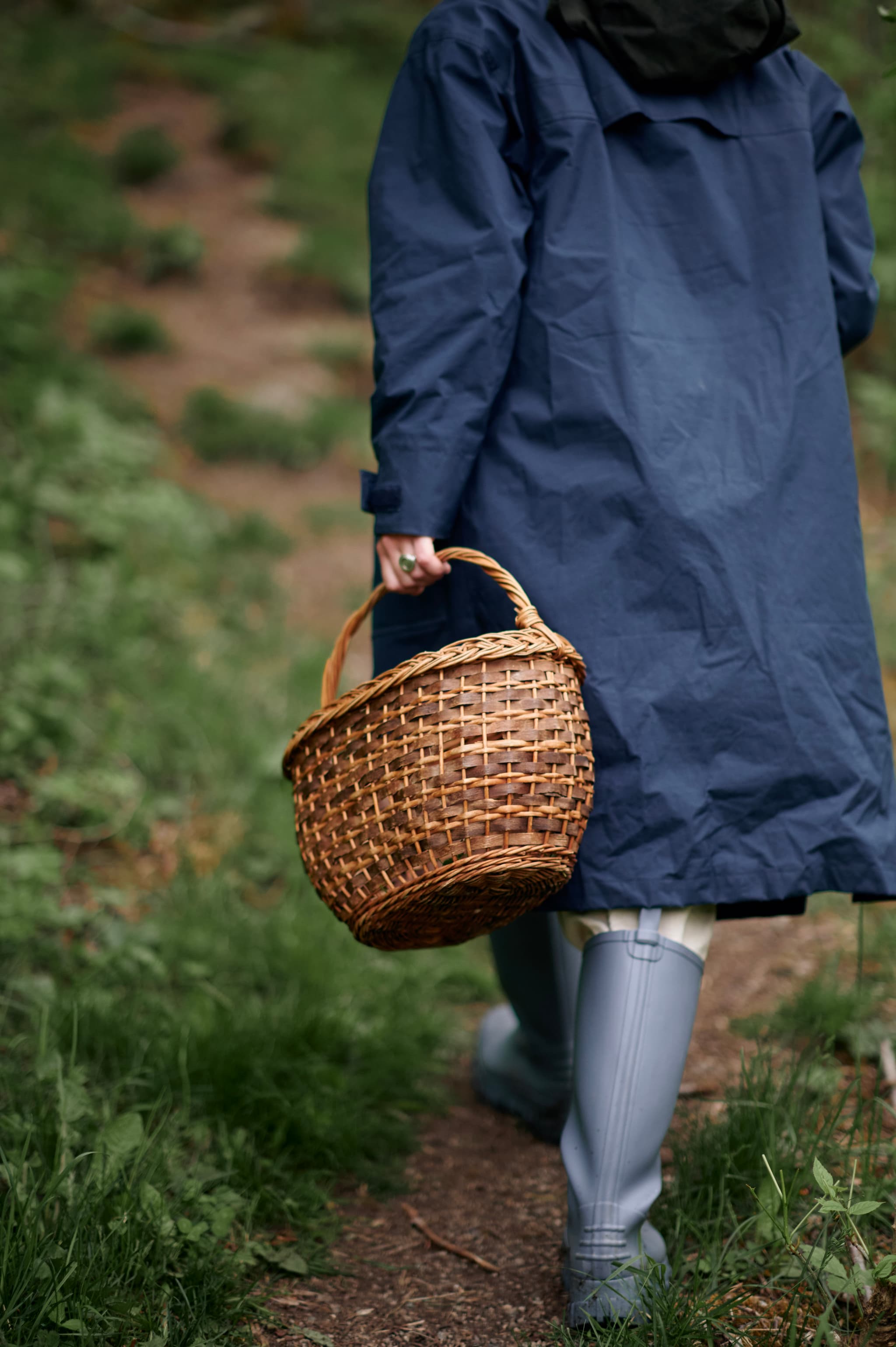 A person in a blue raincoat and rubber boots walks along a grassy path carrying a woven basket.