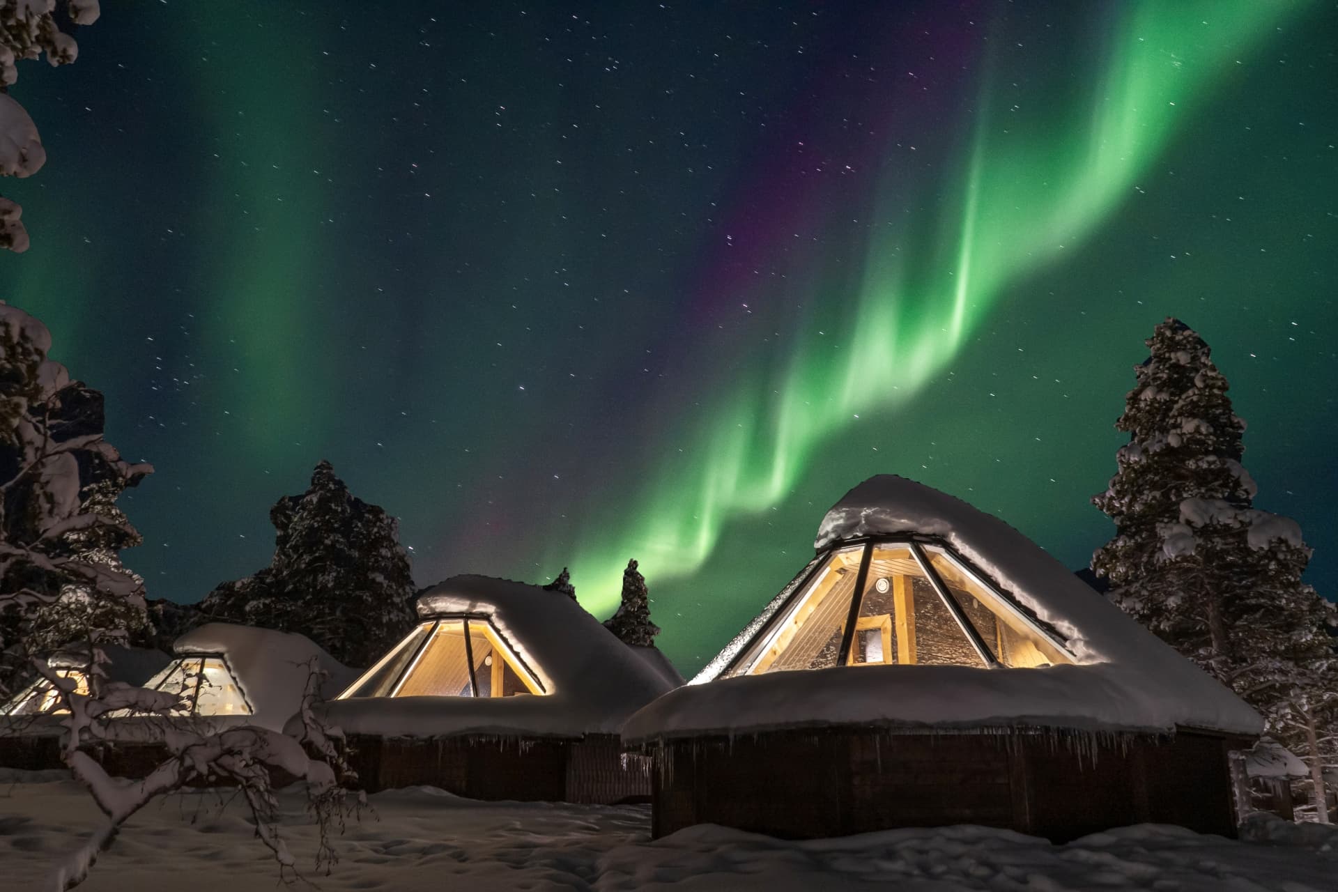 Snow-covered triangular cabins glow warmly beneath a vivid green aurora and starry night sky in a winter forest.