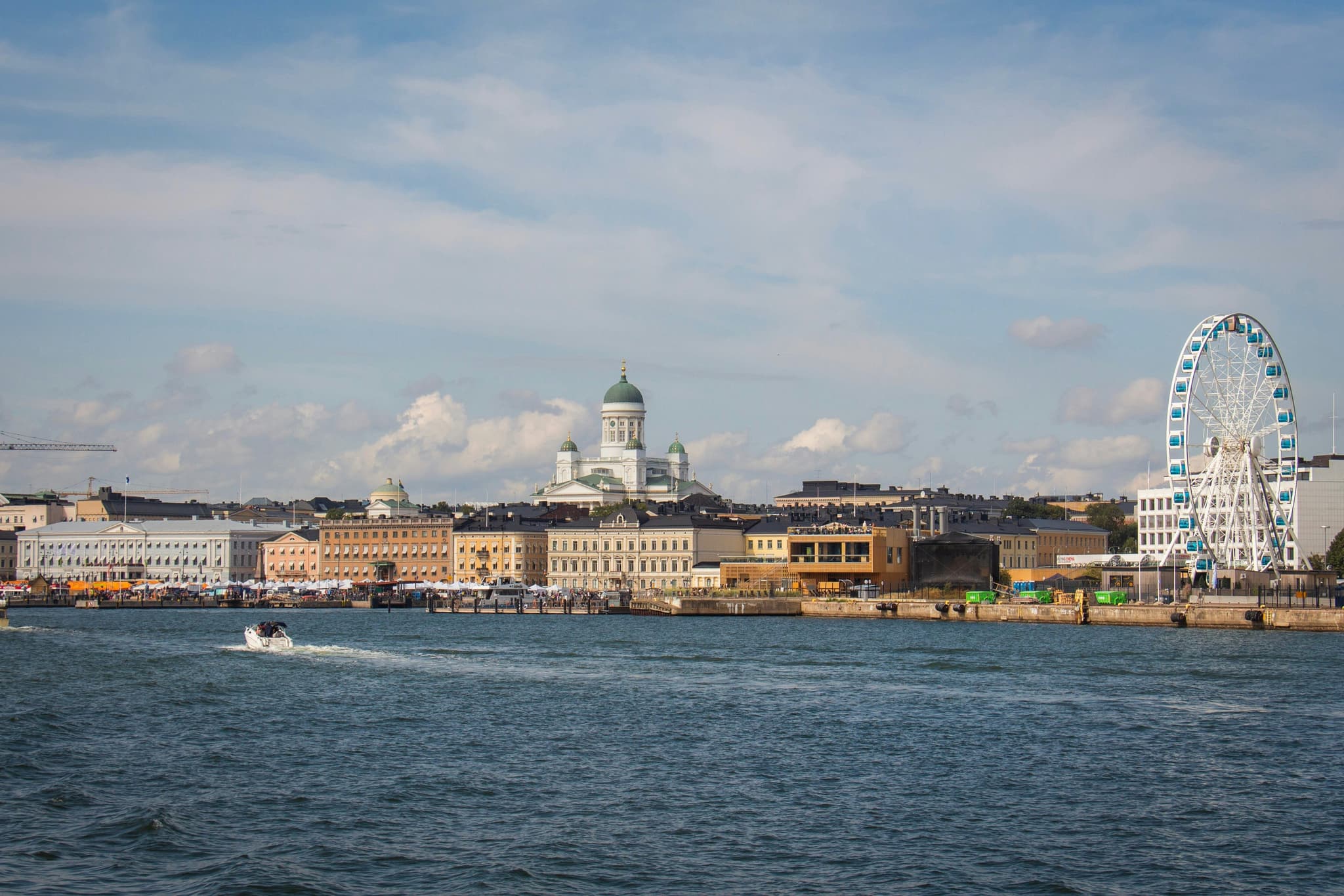 Waterfront city skyline with an opera-house-like domed building and a large Ferris wheel on a waterfront promenade under a partly cloudy sky.