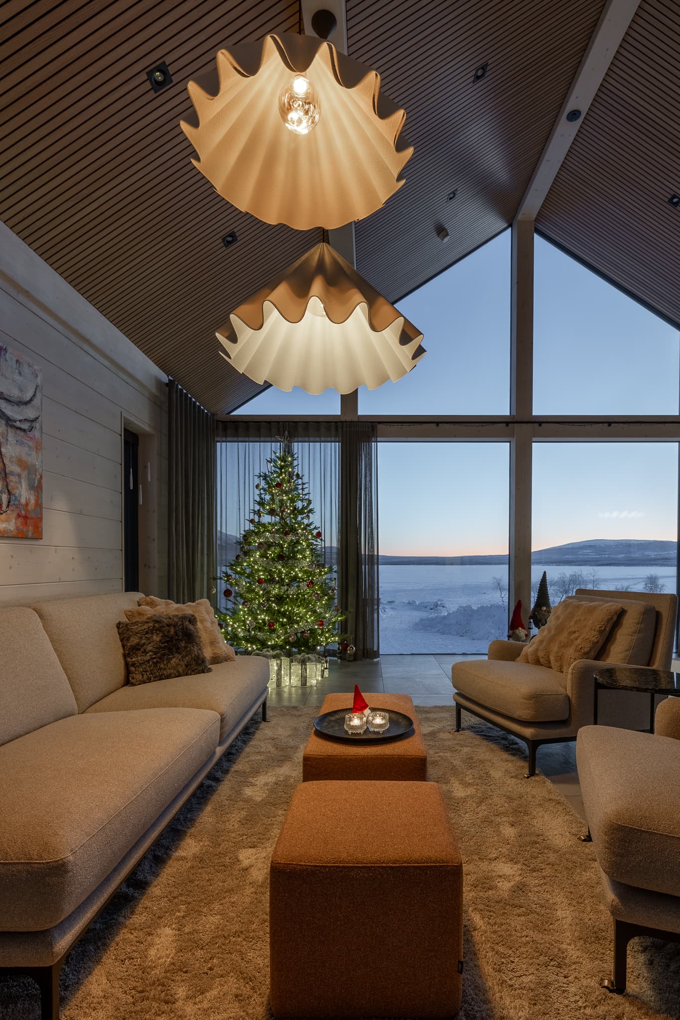 A cozy modern living room with a decorated Christmas tree, two large pendant lights, and floor-to-ceiling windows overlooking a snowy landscape at dusk.