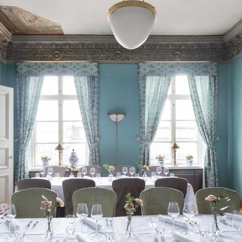 An elegant dining room with set tables, tall windows, patterned curtains, and teal walls.