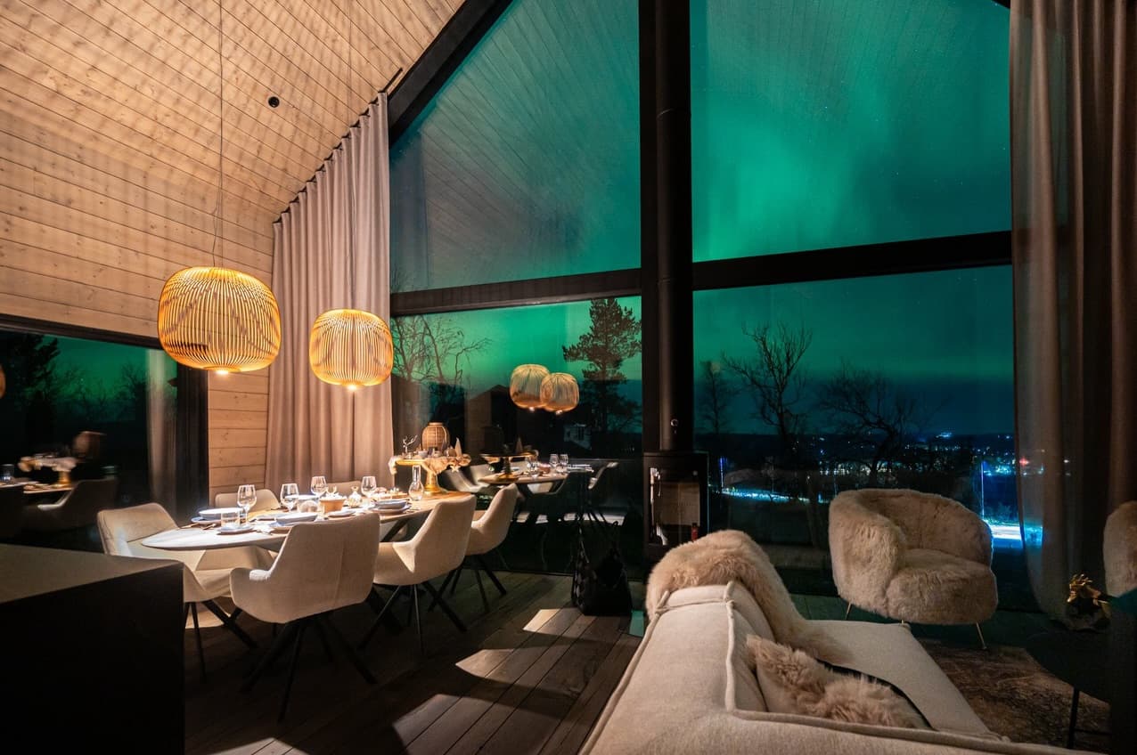 A modern dining room with warmly lit tables and oversized windows overlooking a green night sky.