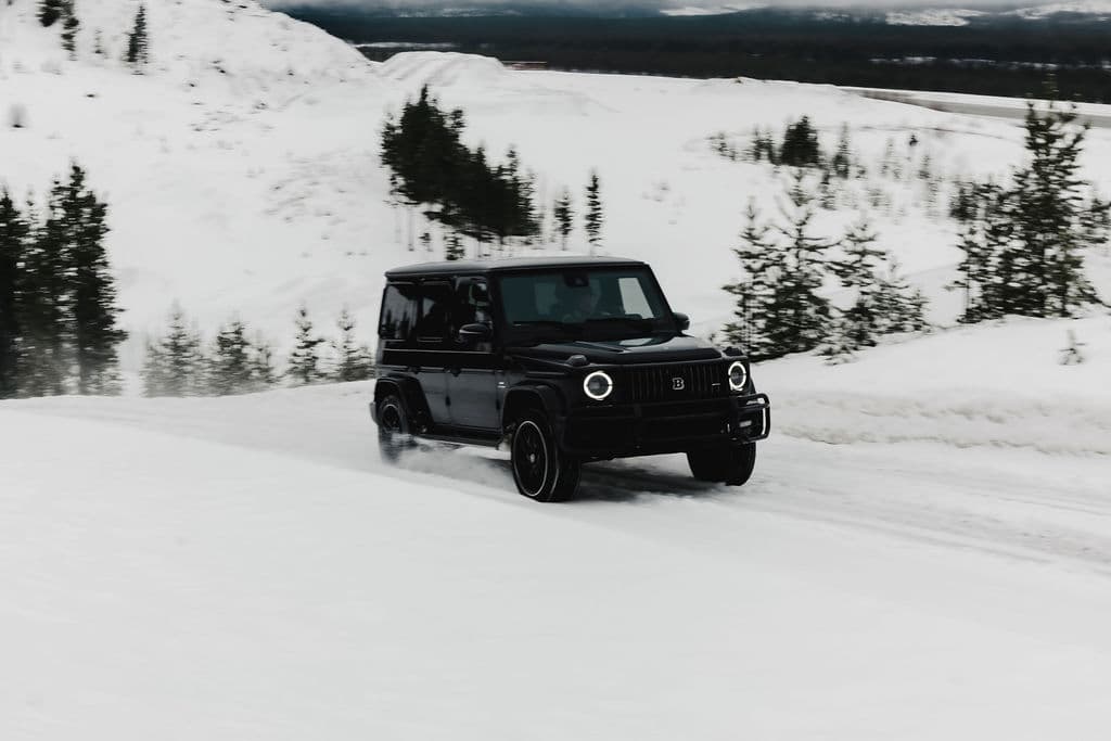 A black off-road SUV drives across a snowy landscape with snow-covered hills and scattered evergreen trees in the background.