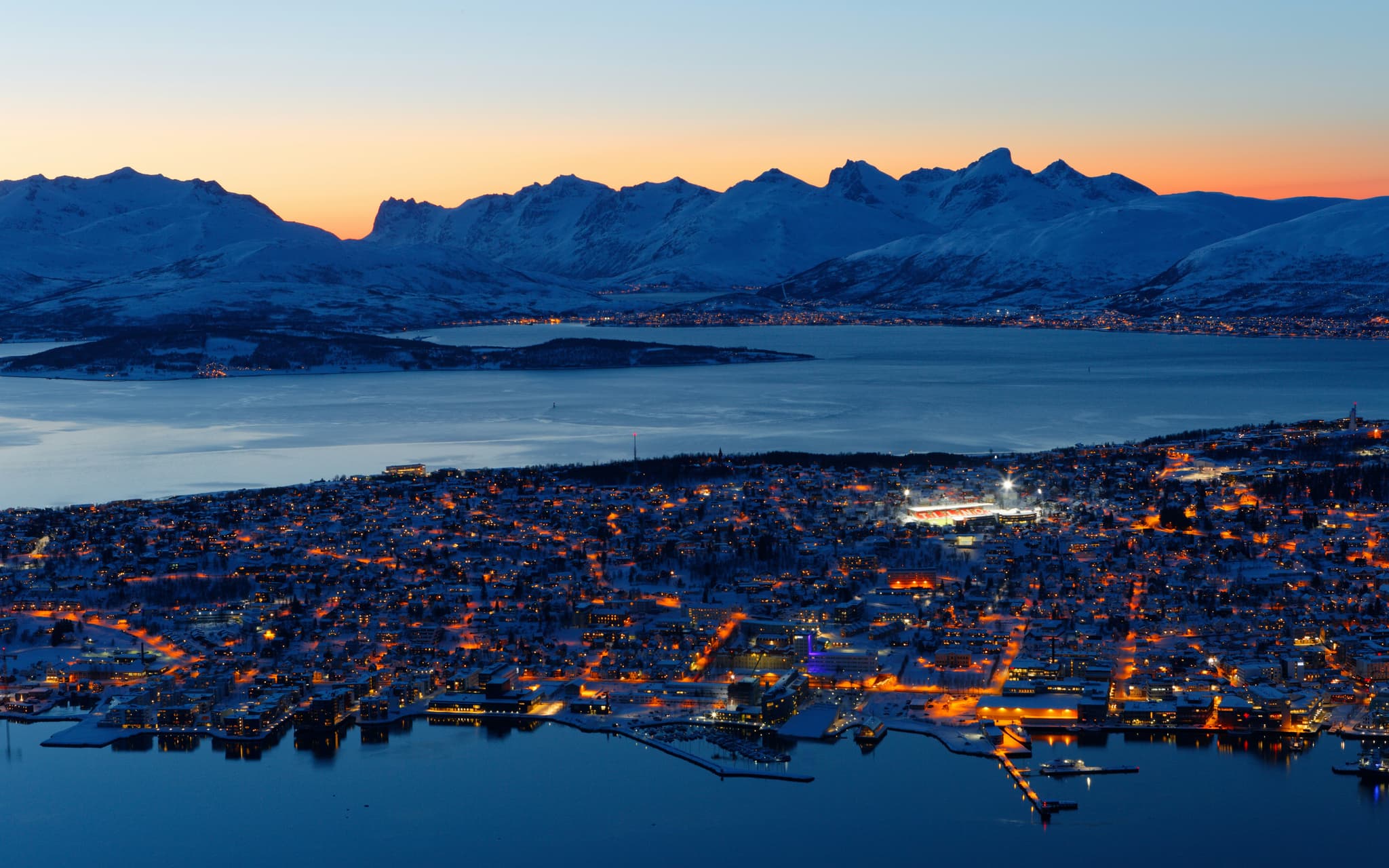 A snowy coastal city glows with orange lights beneath dark mountains at twilight beside a calm fjord.