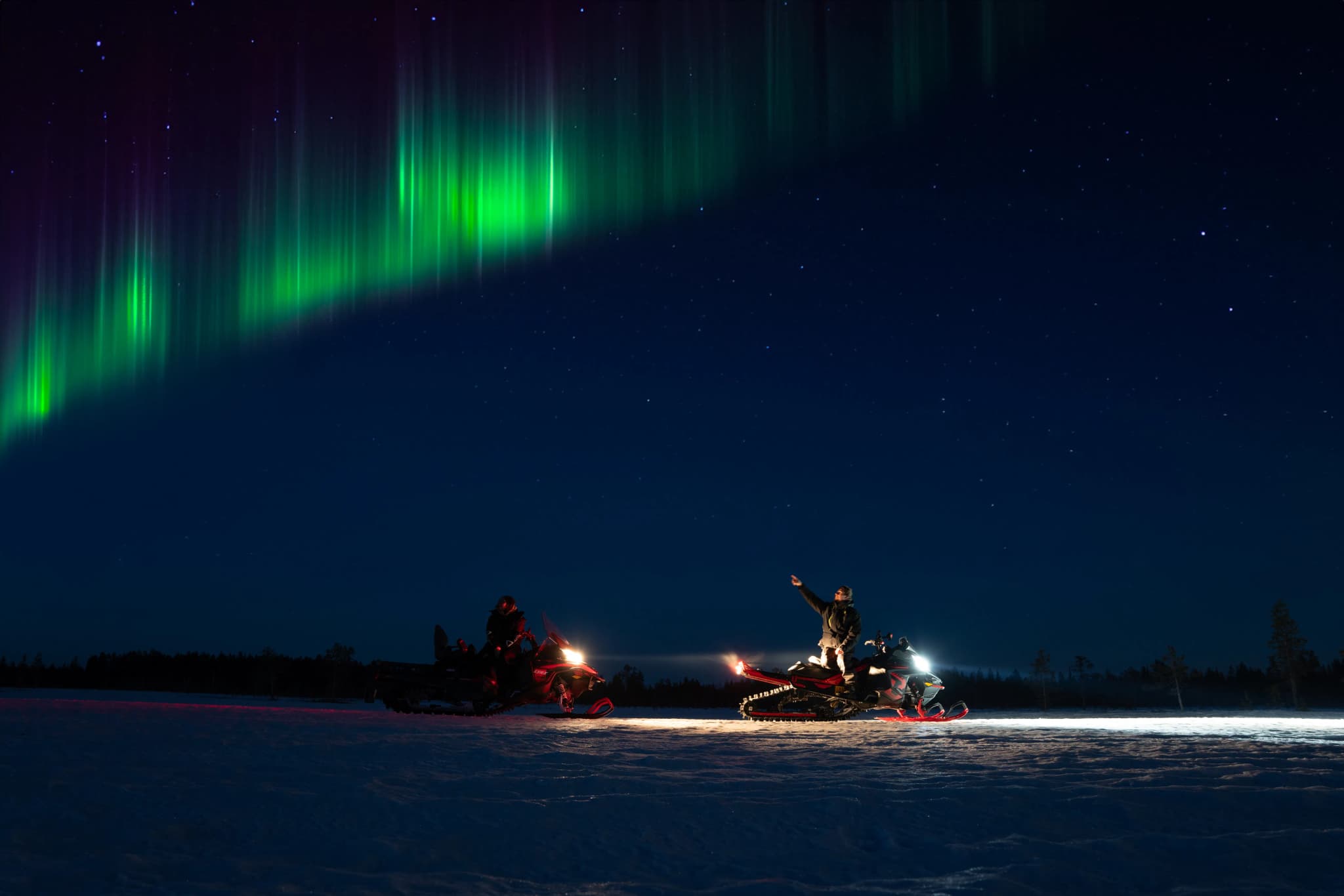 Aurora borealis over a snowy landscape with people riding snowmobiles at night.