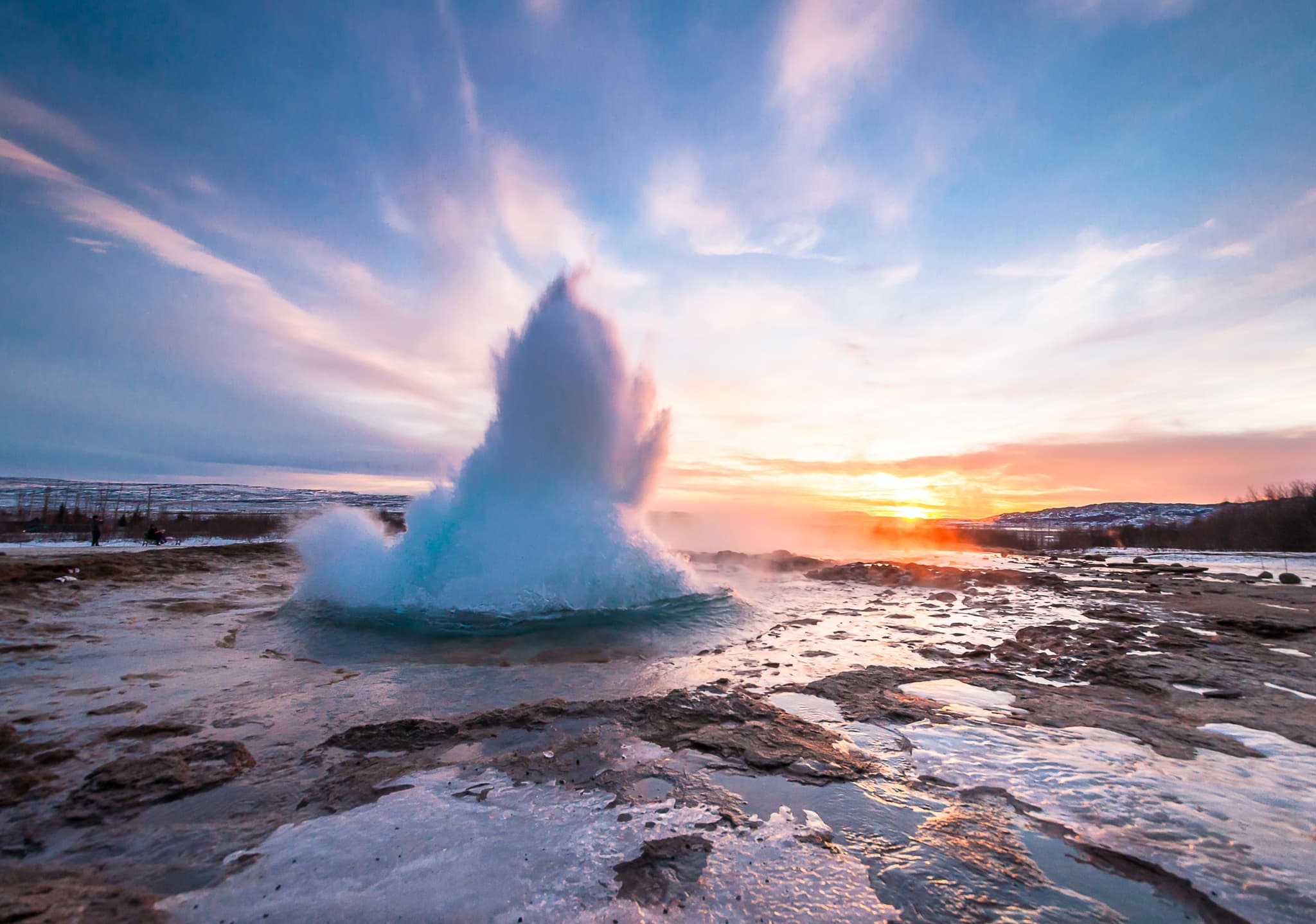 A geyser erupts in a rocky geothermal landscape at sunset under a colorful sky.