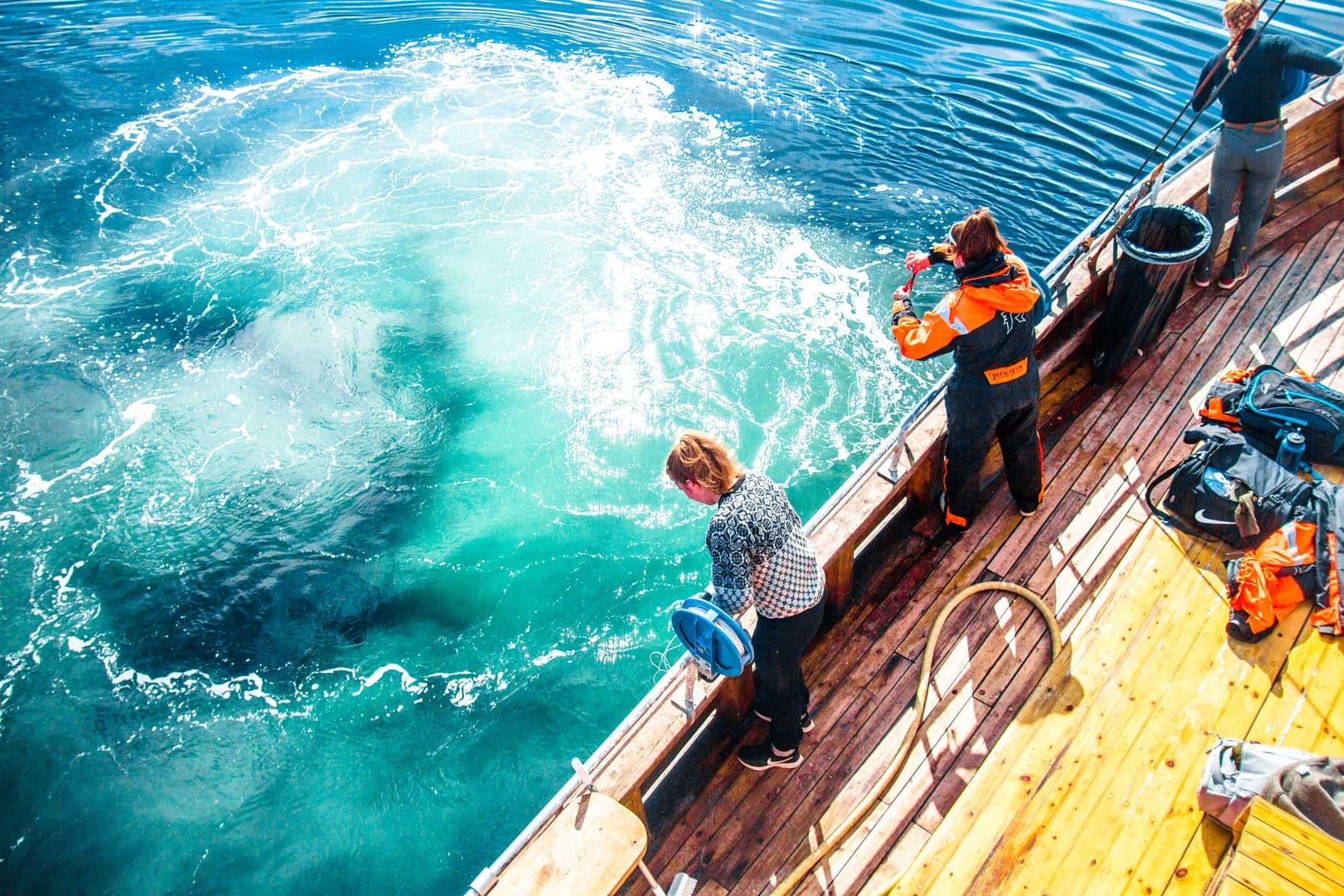 Three people stand on a wooden boat deck while a fishing line disturbs the bright blue water beside the vessel.
