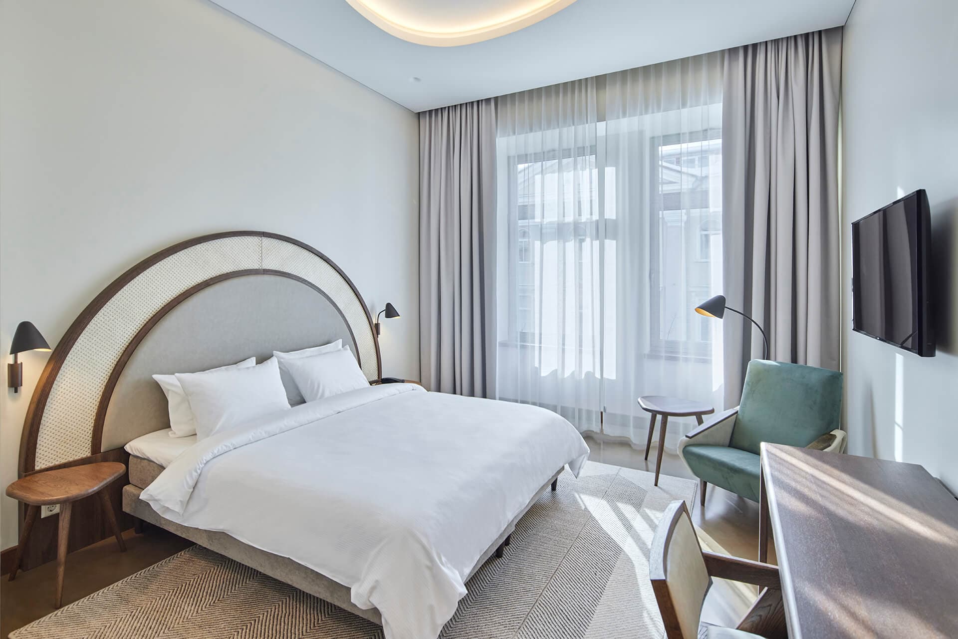 A bright modern hotel bedroom features a double bed with white linens, a curved upholstered headboard, a desk chair, and large windows with sheer curtains.