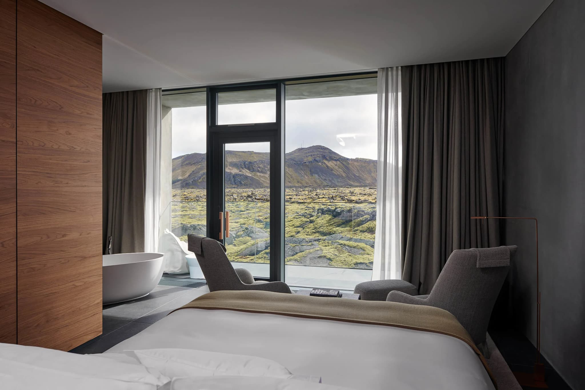 Modern bedroom with floor-to-ceiling windows and mountain landscape view, gray curtains, and a freestanding bathtub