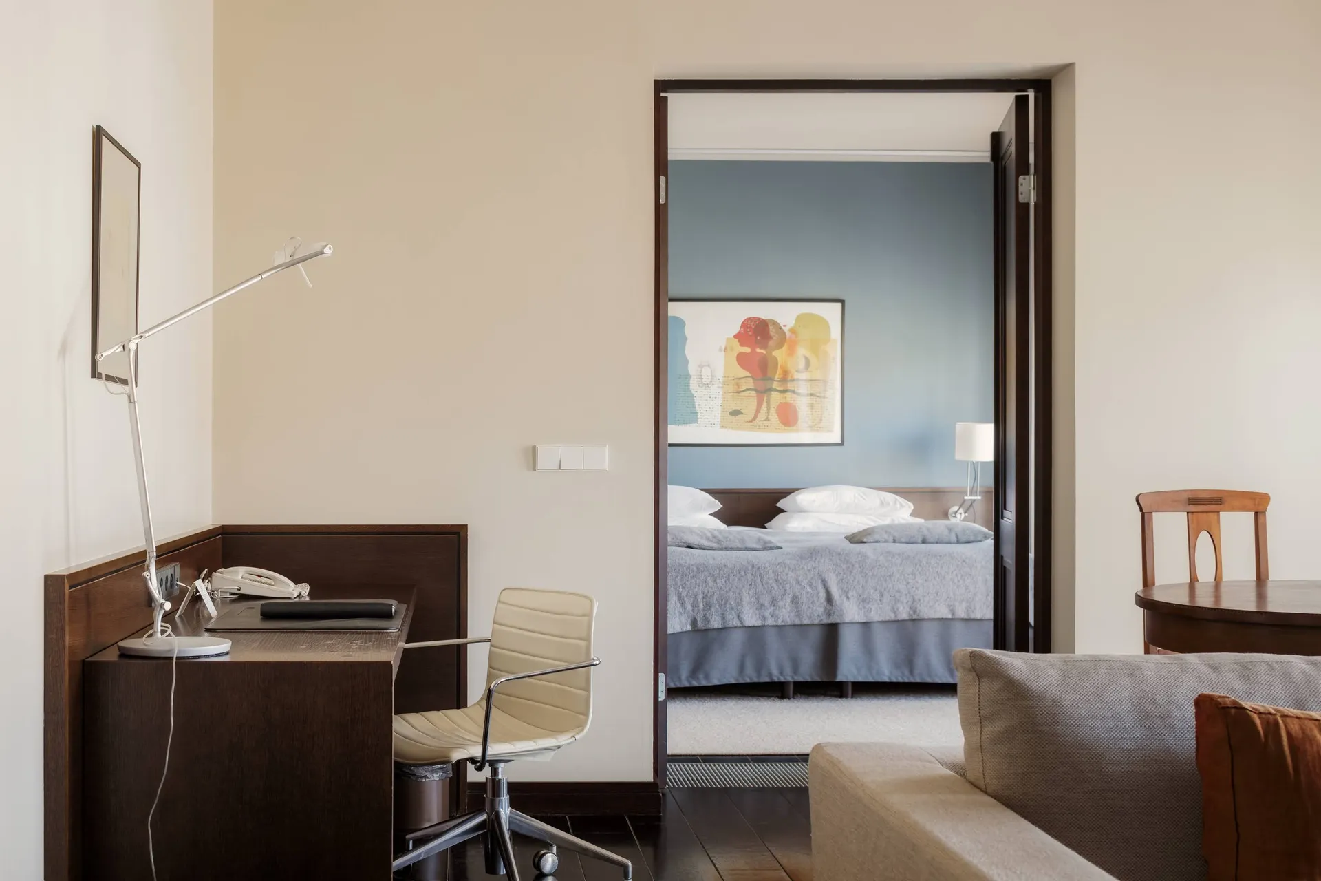 A modern apartment-style hotel suite shows a desk and chair in the foreground with a bedroom visible through an open doorway.