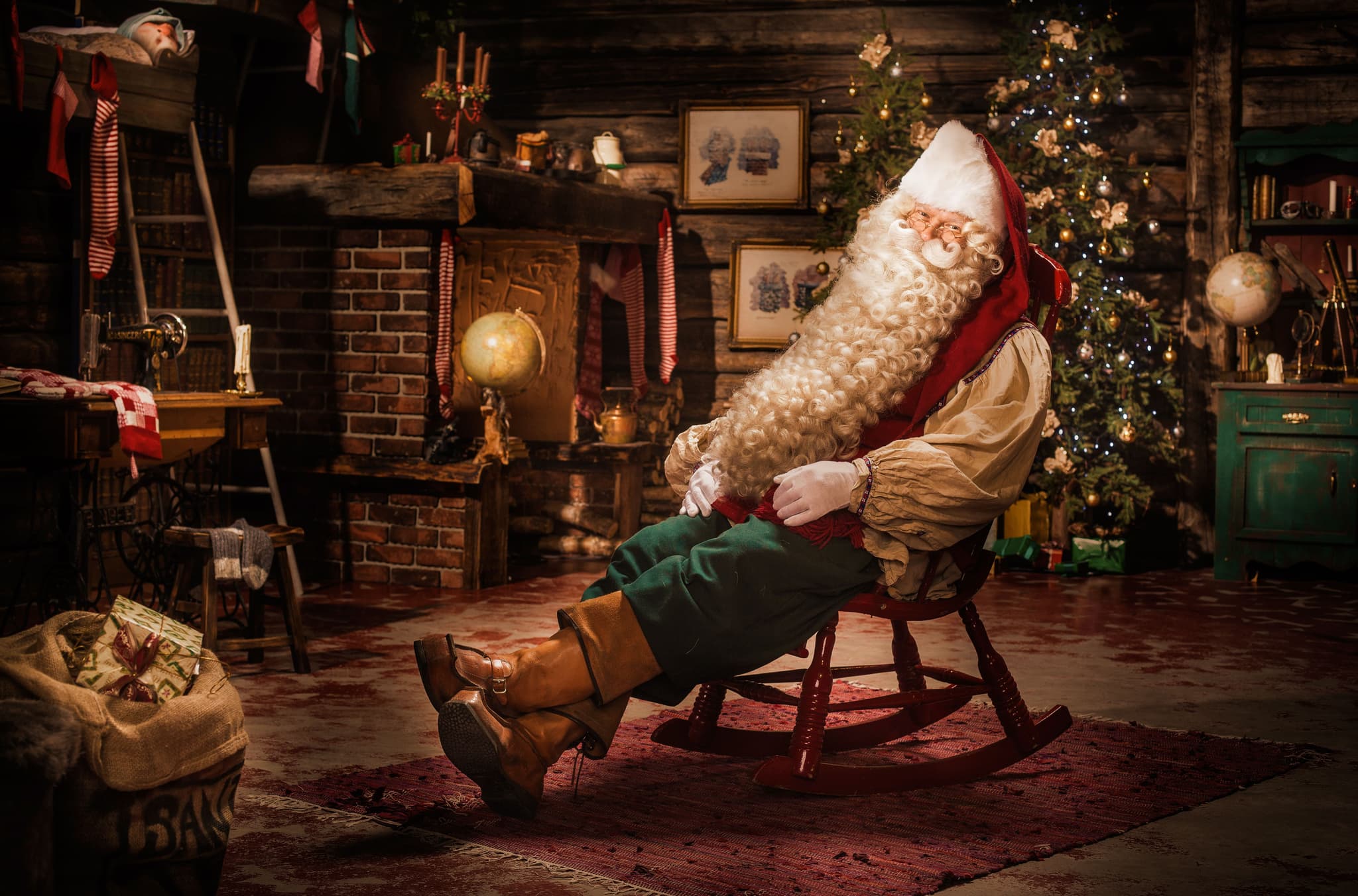 Santa Claus sits in a cozy rustic room decorated with Christmas trees, gifts, and holiday ornaments.