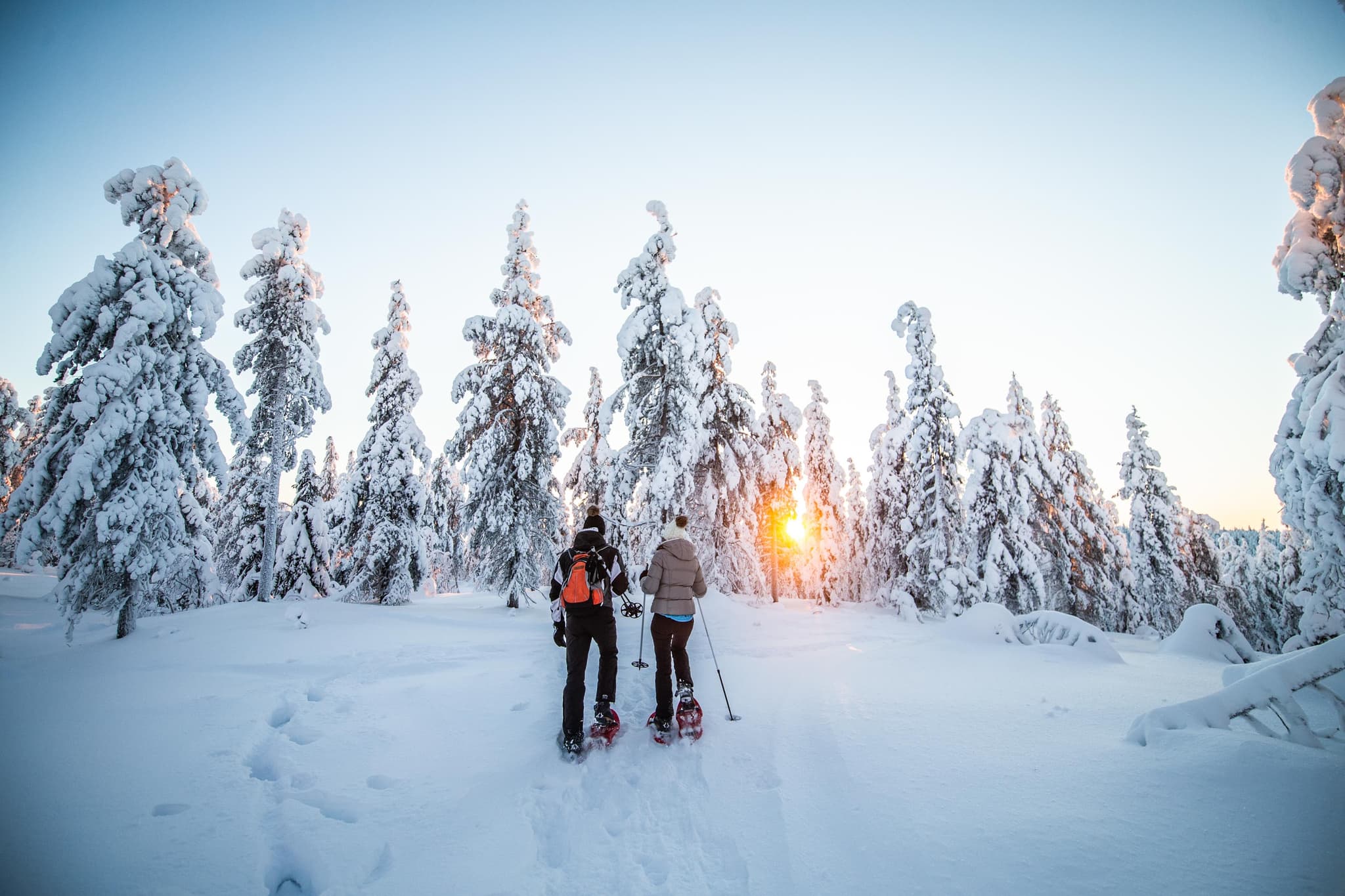 Two snowshoers walking on a snowy trail at sunrise with frost-covered pine trees and low winter sun behind the landscape.