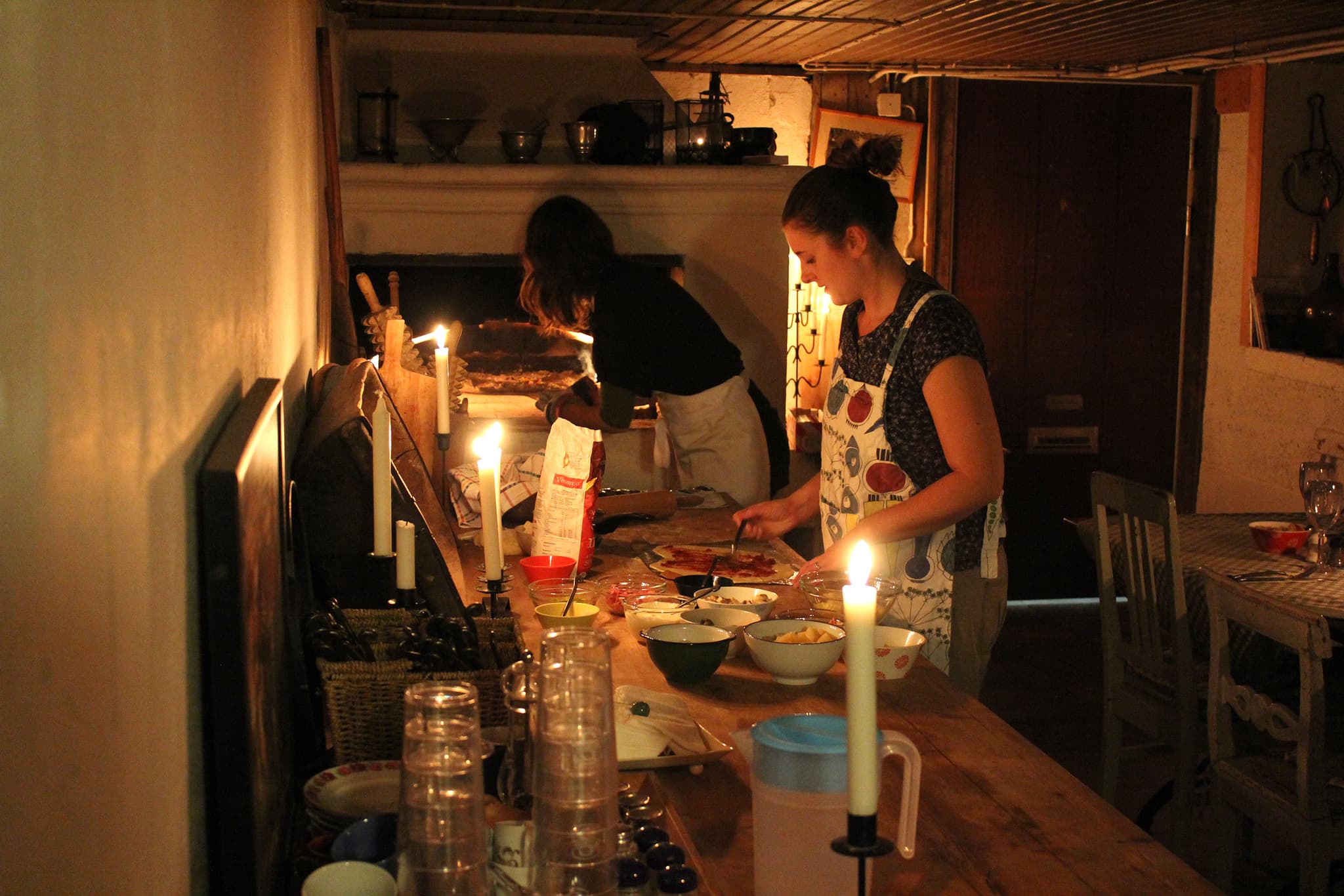 Warm candlelit dinner scene inside a rustic room with people preparing and baking pizza in an outdoor-style brick oven.