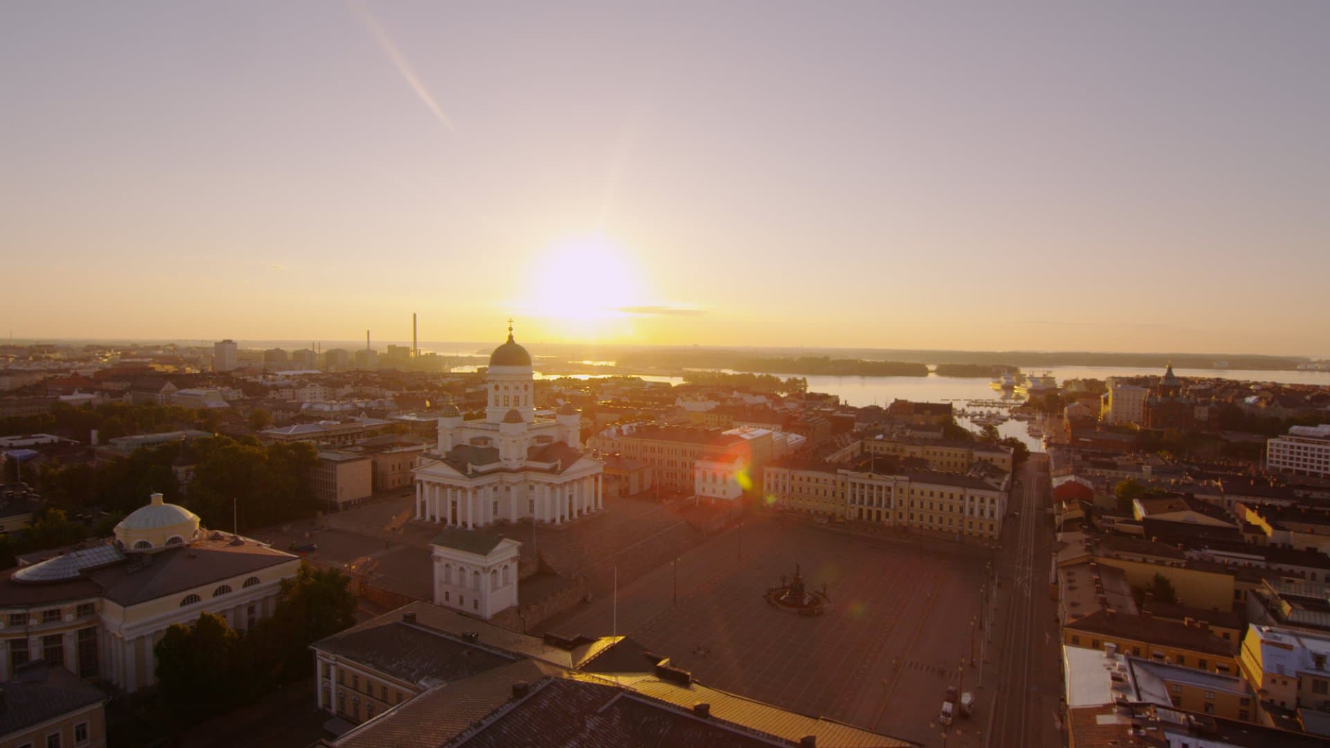 A golden sunset lights up a city square with a white cathedral and waterfront skyline in the distance.