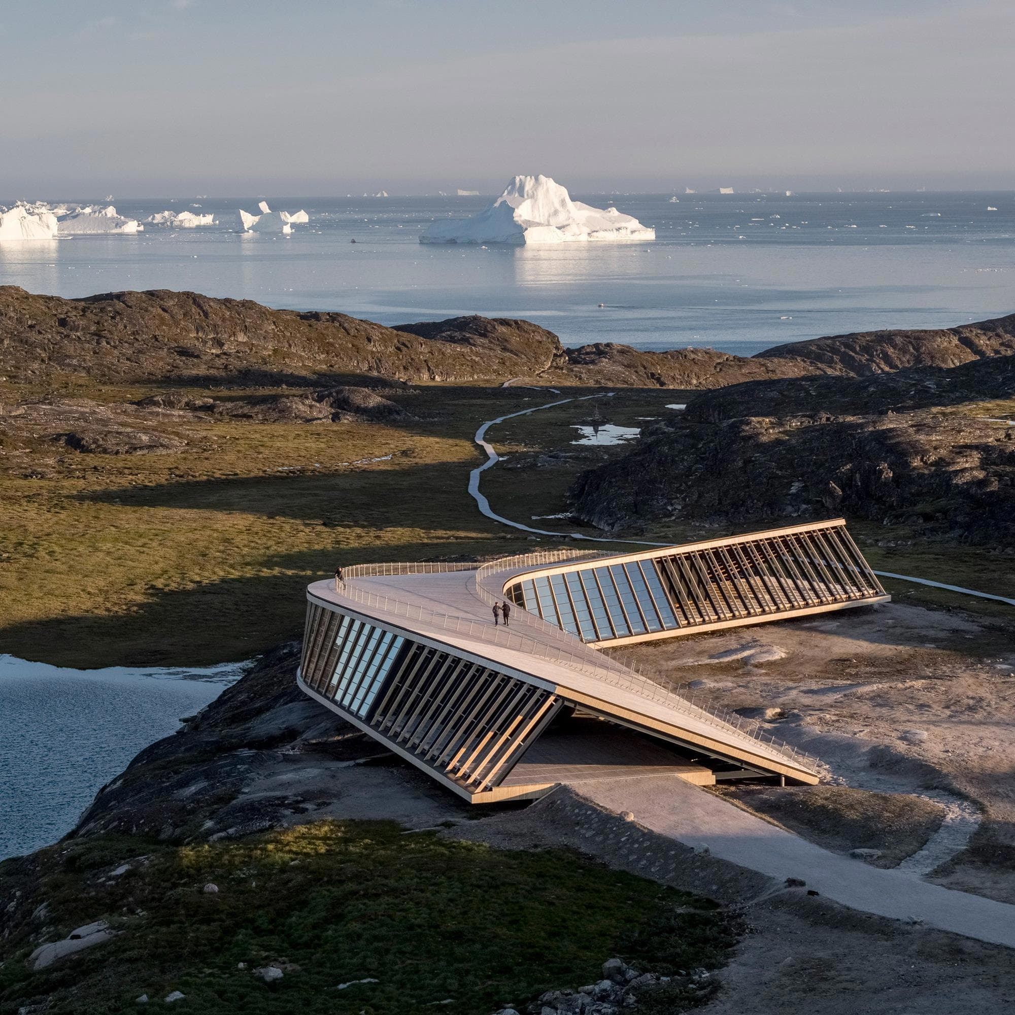 A modern angular building sits on a rocky Greenland coastline overlooking calm water with icebergs in the distance.