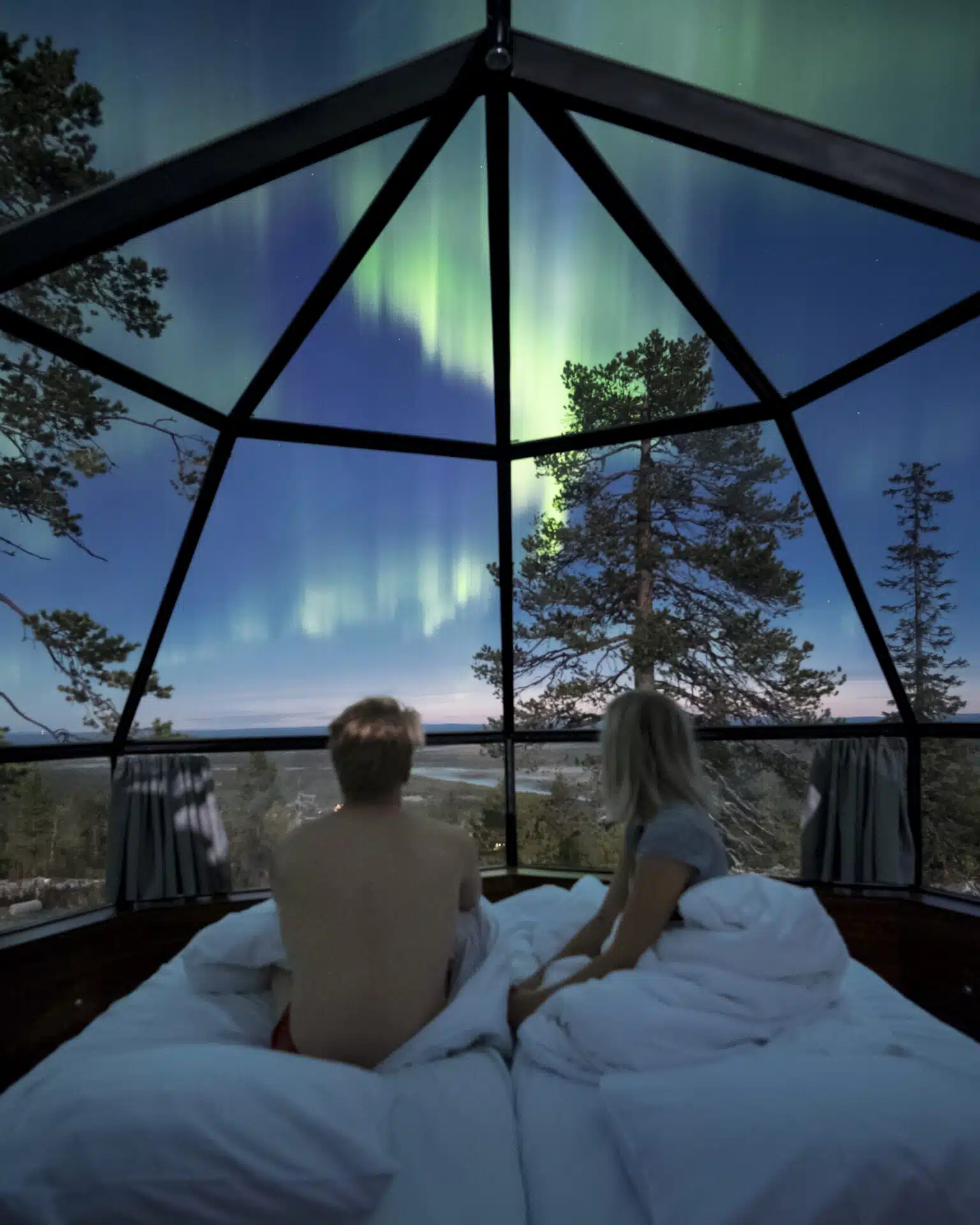 Two people sit up in a glass igloo bed and look out at the northern lights over a snowy forest landscape at dusk.
