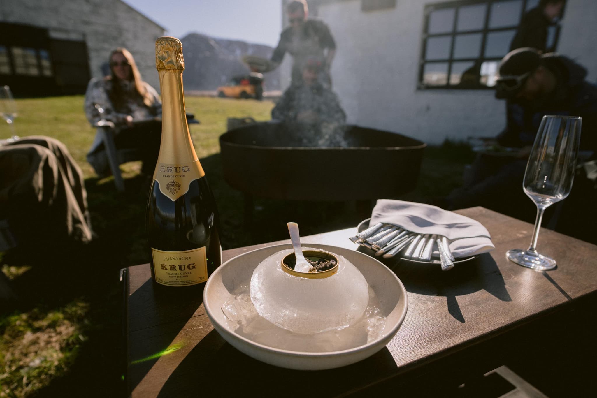A bottle of champagne, a wine glass, and a small table setting with an ice bowl sit outdoors beside people gathered around a fire pit.
