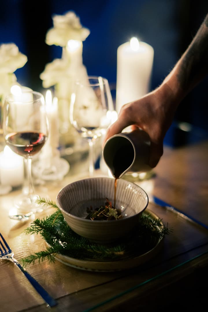 A hand pours sauce into a bowl at a candlelit table set with wine glasses and evergreen decorations.