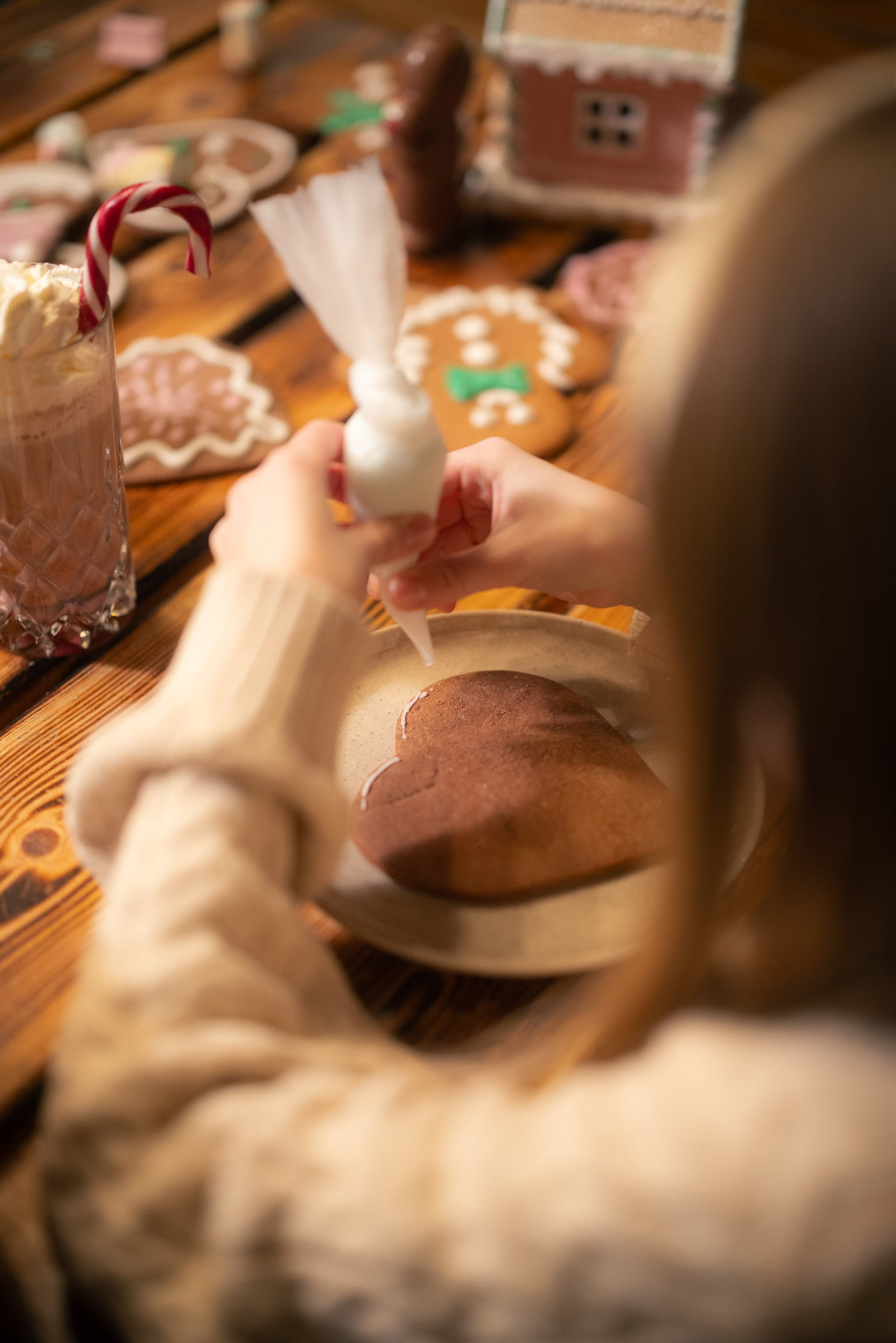 A child decorates a gingerbread heart with icing at a wooden table surrounded by holiday cookies and candy canes.