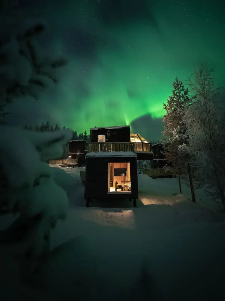 Snow-covered modern cabins glow warmly beneath a vivid green aurora in a dark winter landscape.