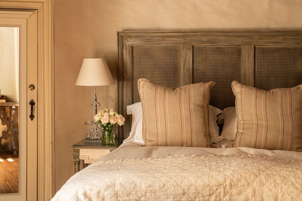 A warmly lit bedroom shows a neatly made bed with striped pillows, a quilted coverlet, a bedside lamp, and a small vase of flowers beside the headboard.