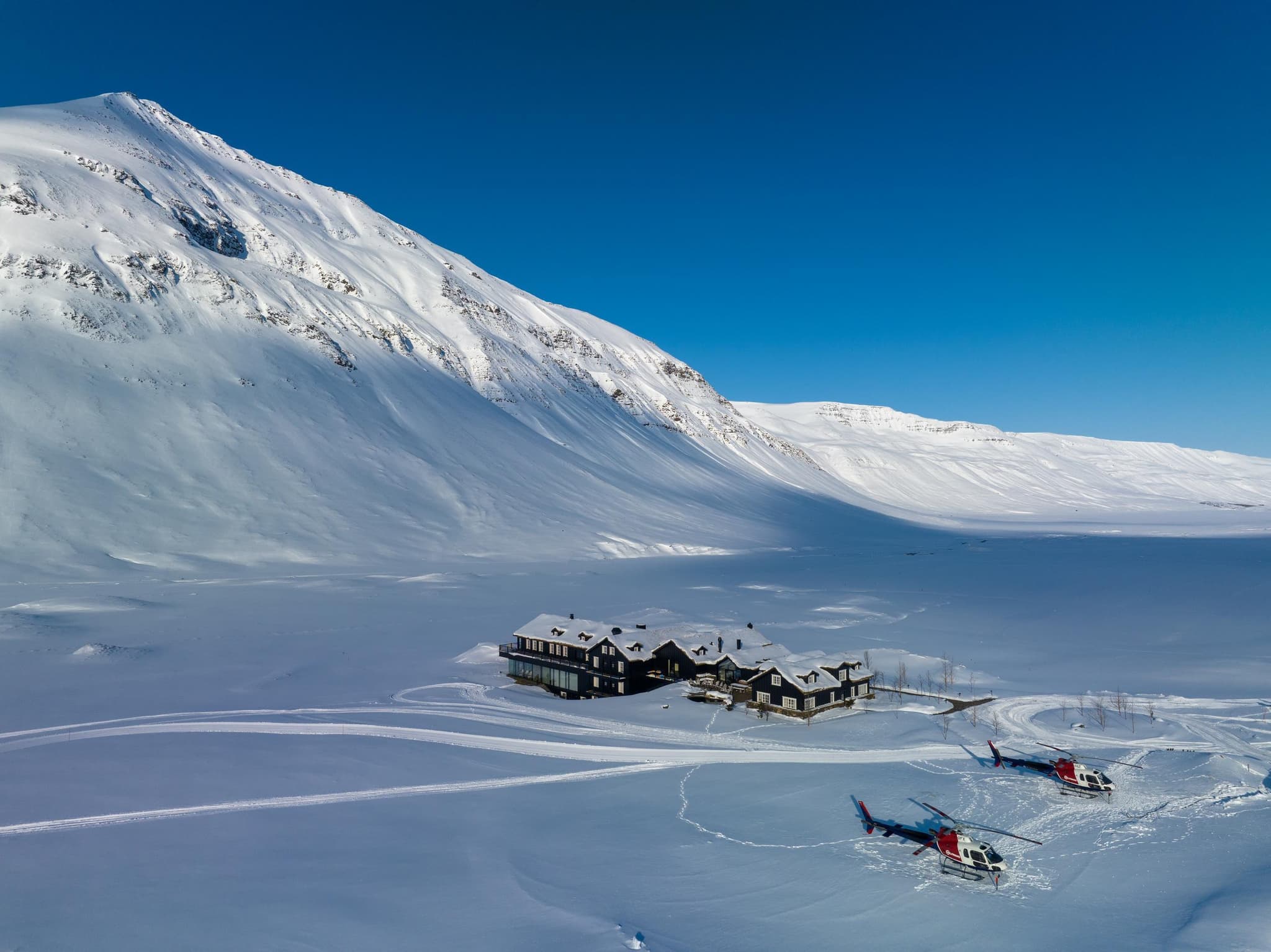 A snow-covered lodge sits in a wide mountain valley beneath steep white slopes and a clear blue sky, with two helicopters parked in the foreground.