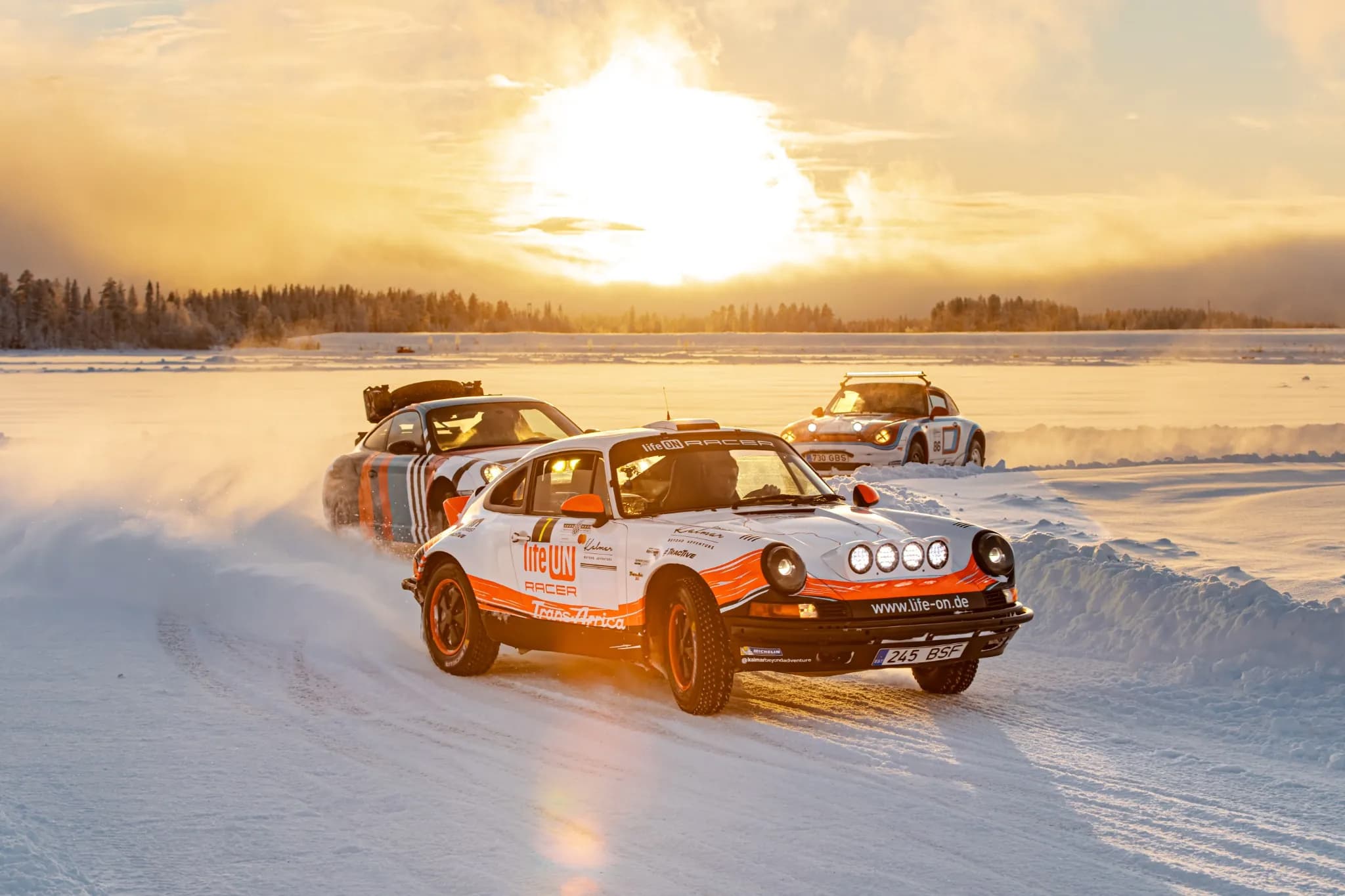 Three rally cars drive across a snowy landscape at sunset, kicking up snow under a bright golden sky.